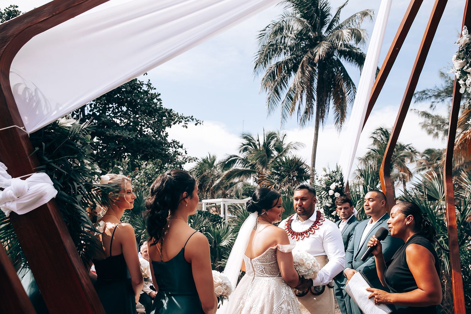 Wedding guests seated under a white canopy with palm trees visible in the background.
