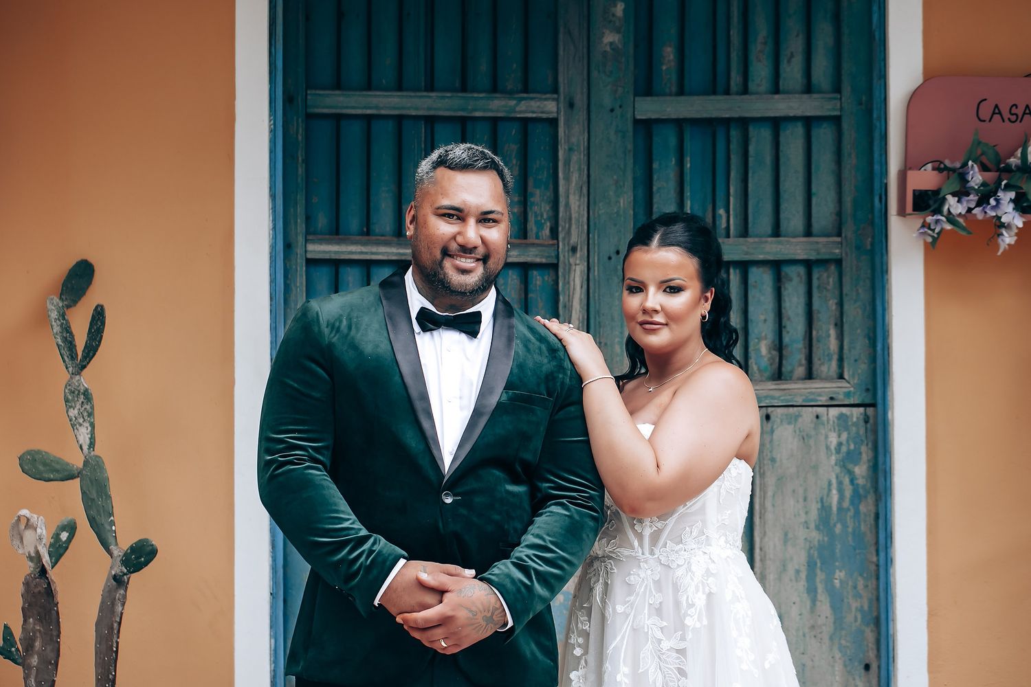A couple poses together in formal wedding attire in front of a rustic blue door and peach-colored wall.