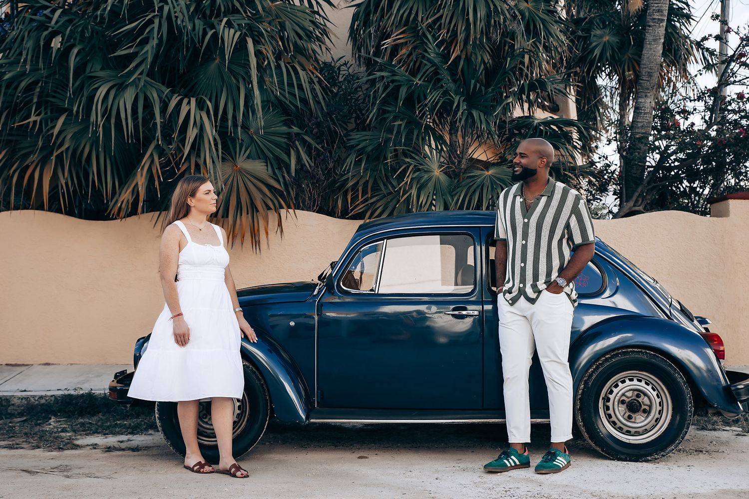 Interracial couple posing beside a classic blue Volkswagen Beetle with tropical palm trees background.