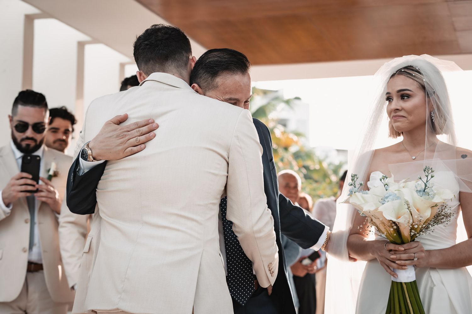 Emotional groom embracing father at the altar as the bride watches with bouquet in hand.