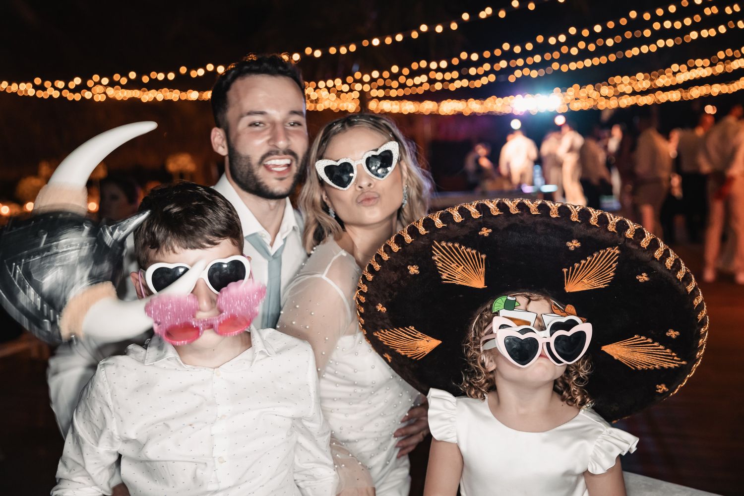 Wedding couple posing with two children wearing fun novelty sunglasses and a sombrero at reception.