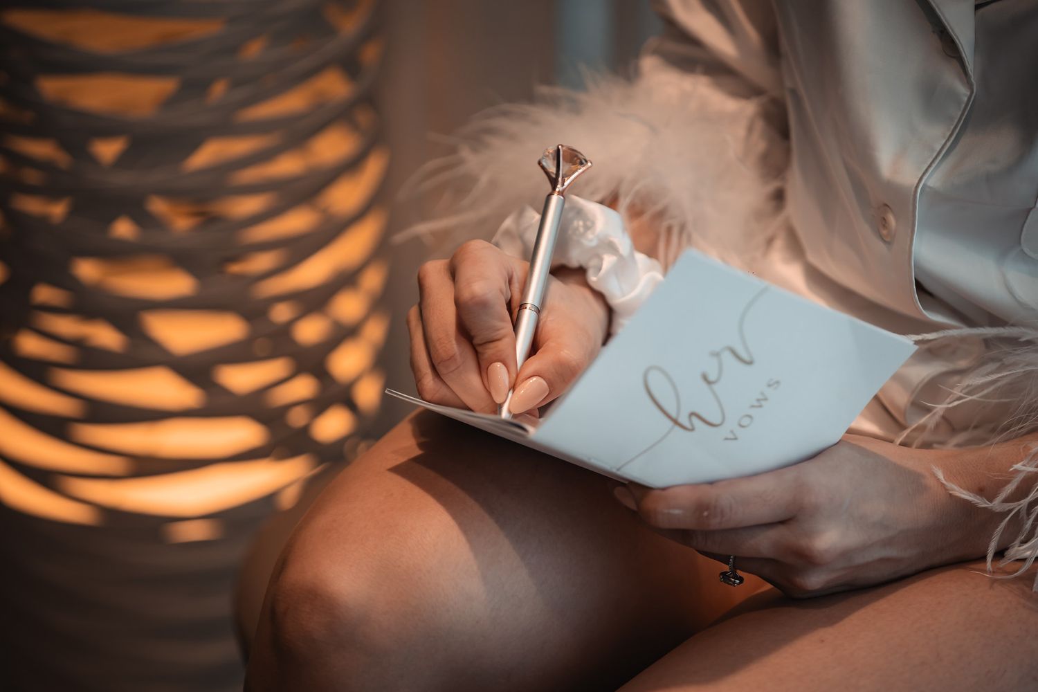 A bride writes in a white journal with a feather pen beside a glowing decorative lamp.