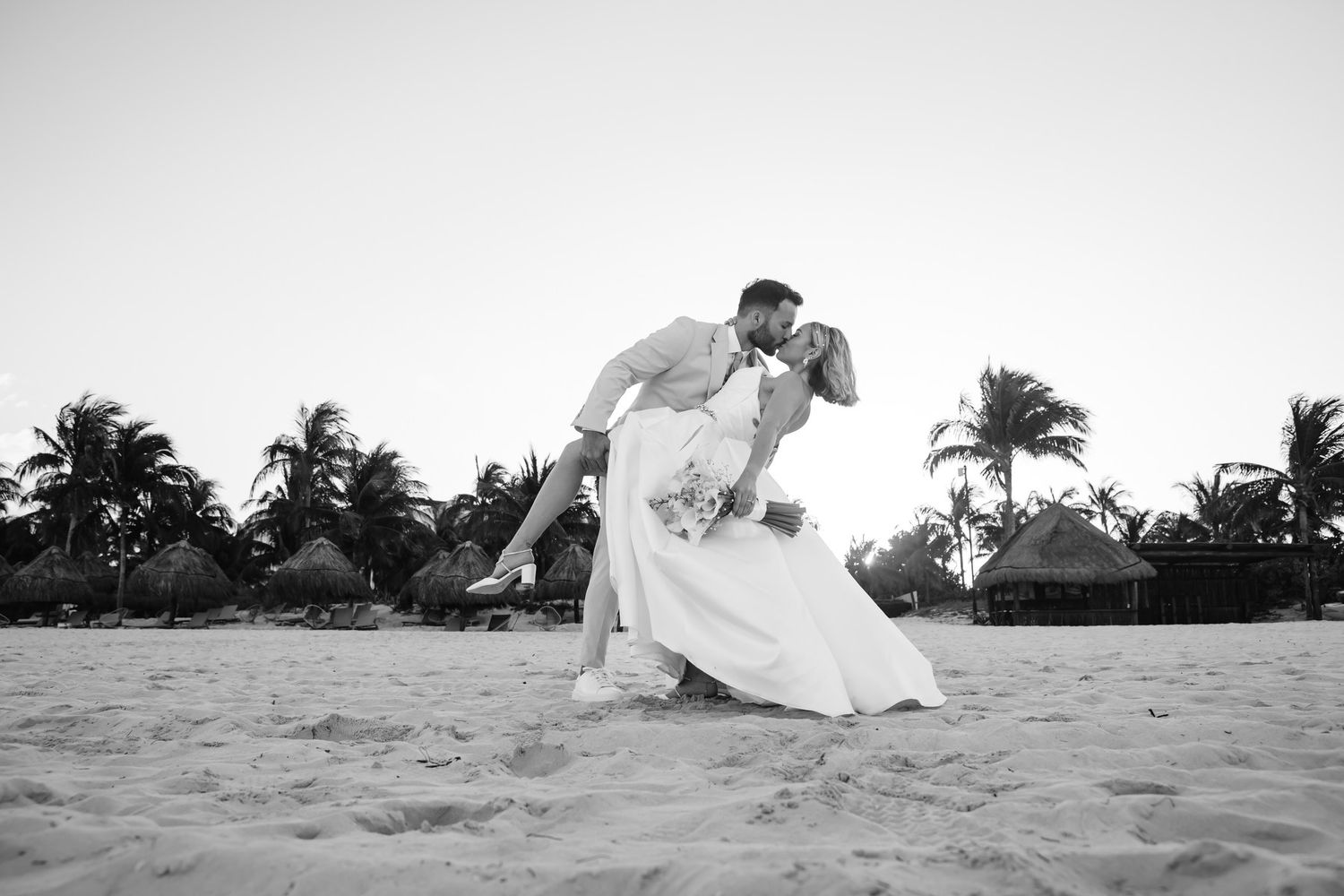 Bride and groom share a romantic dip kiss on a sandy beach with palm trees in black and white.