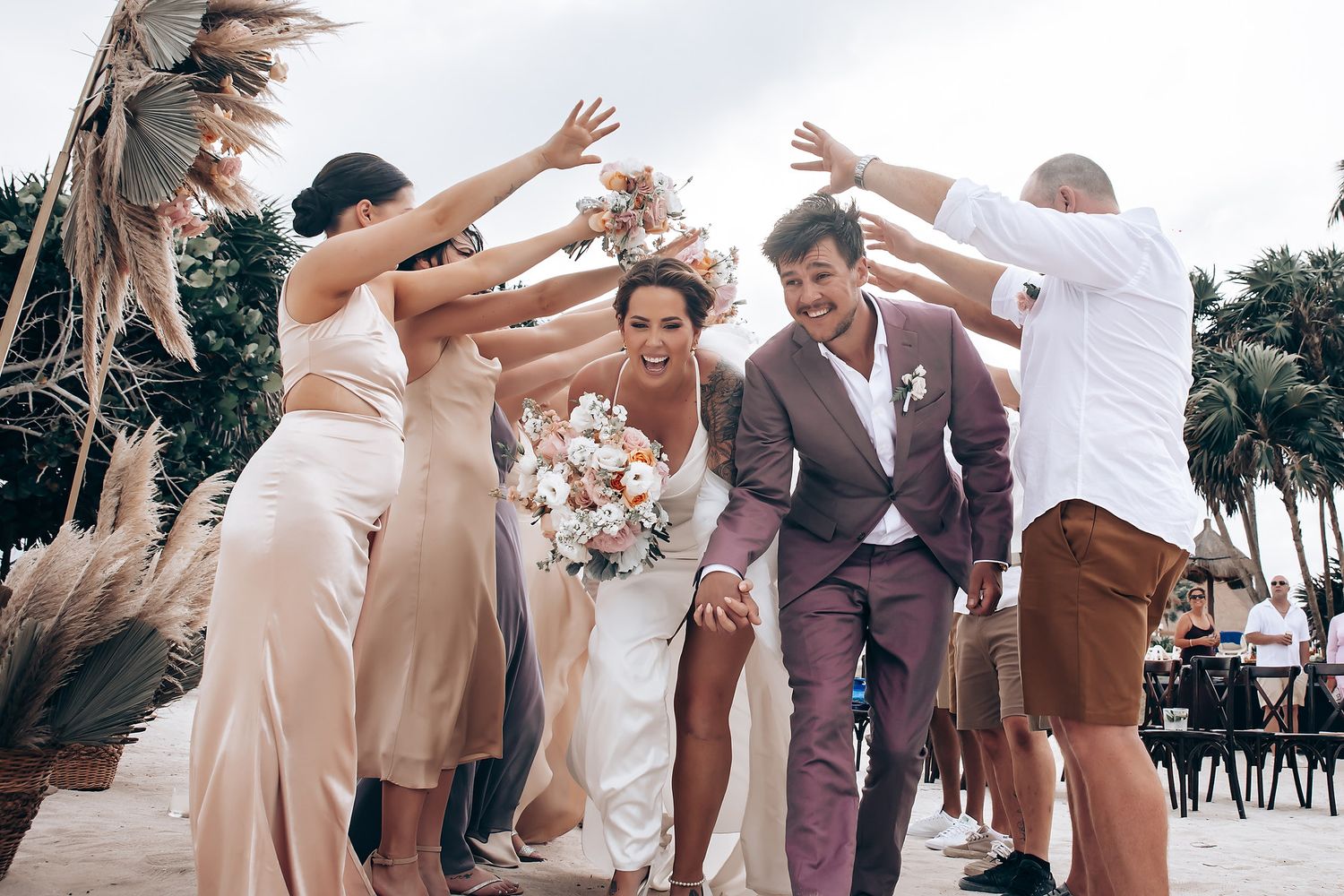 Wedding celebration with guests in beige and neutral colors dancing outdoors near palm trees.
