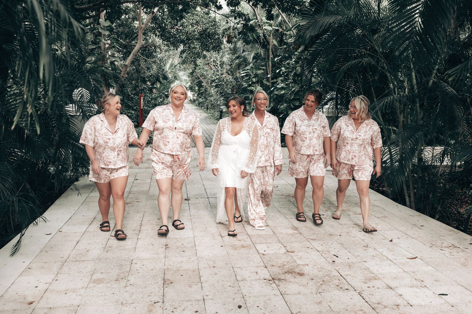 Bride and bridesmaids walking together in matching floral pajama sets through tropical garden path.