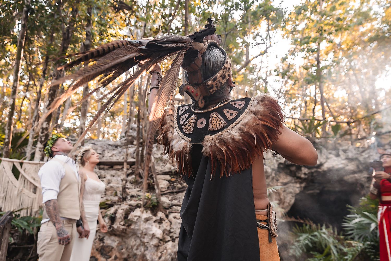 Mayan shaman in traditional headdress and feathered costume performs ceremonial ritual outdoors in jungle ruins setting.