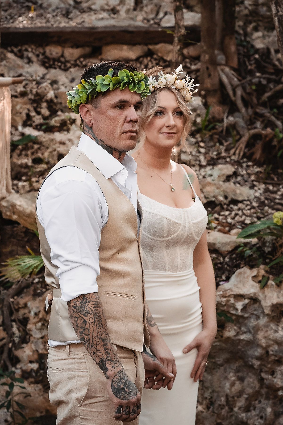 Tattooed groom in tan vest and bride in white dress wear floral crowns at an outdoor bohemian wedding.
