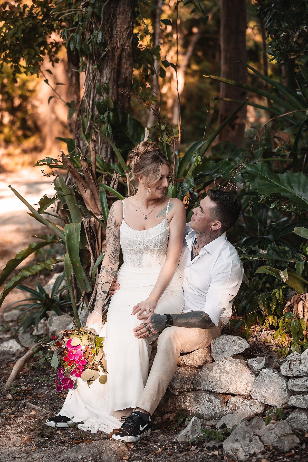 Bride and groom sitting on stone wall surrounded by tropical greenery, sharing a romantic moment on their wedding day.
