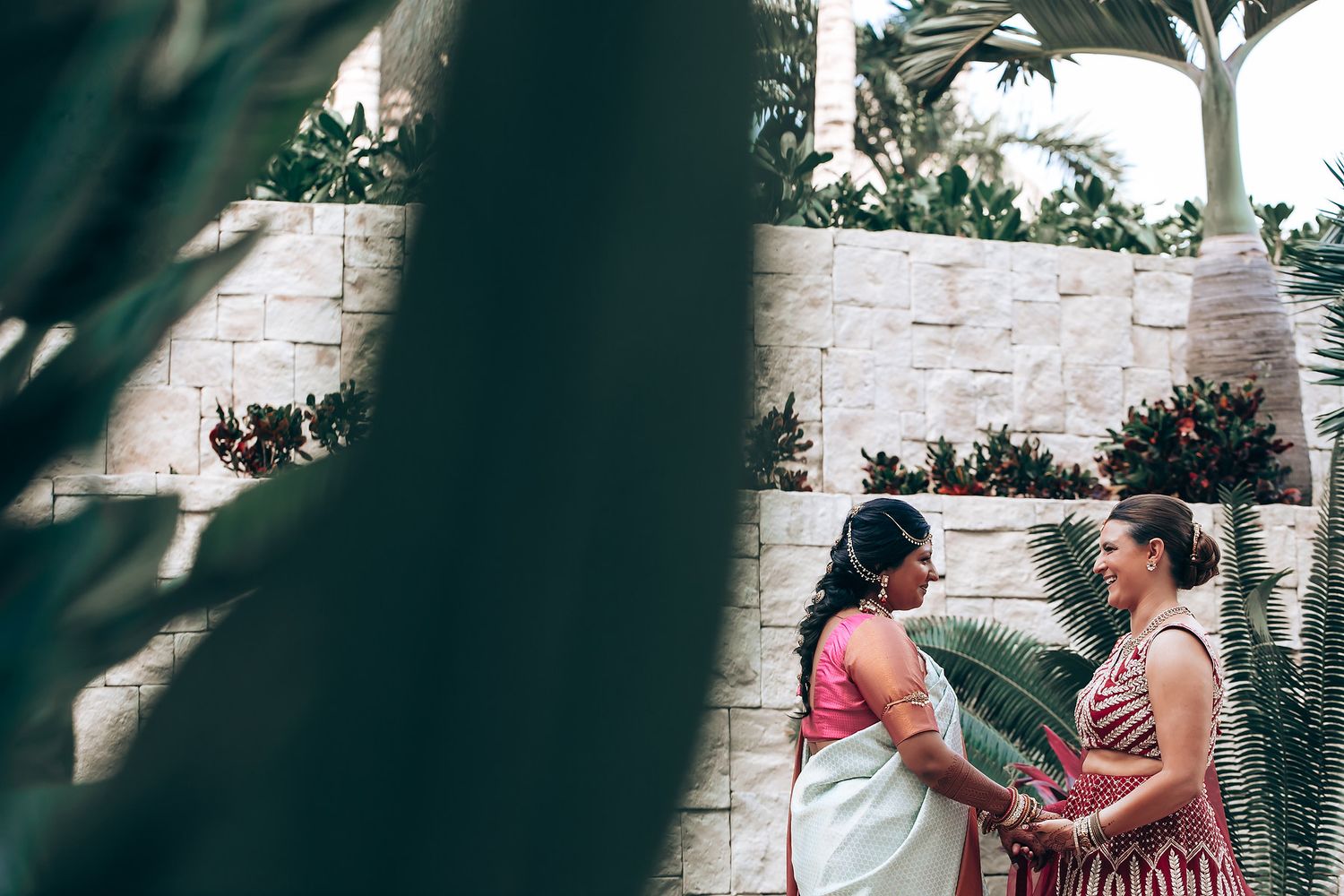 Intimate moment captured through foliage at a tropical wedding venue with stone walls.