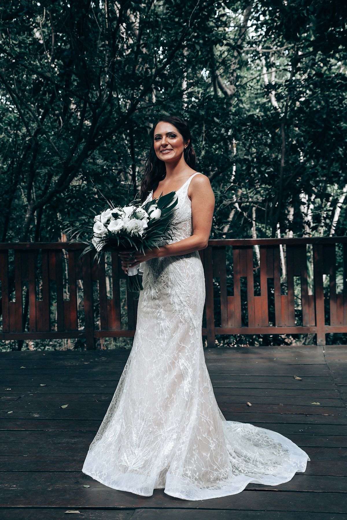 Elegant lace wedding dress with a flowing train and white bouquet photographed on a wooden deck surrounded by greenery.