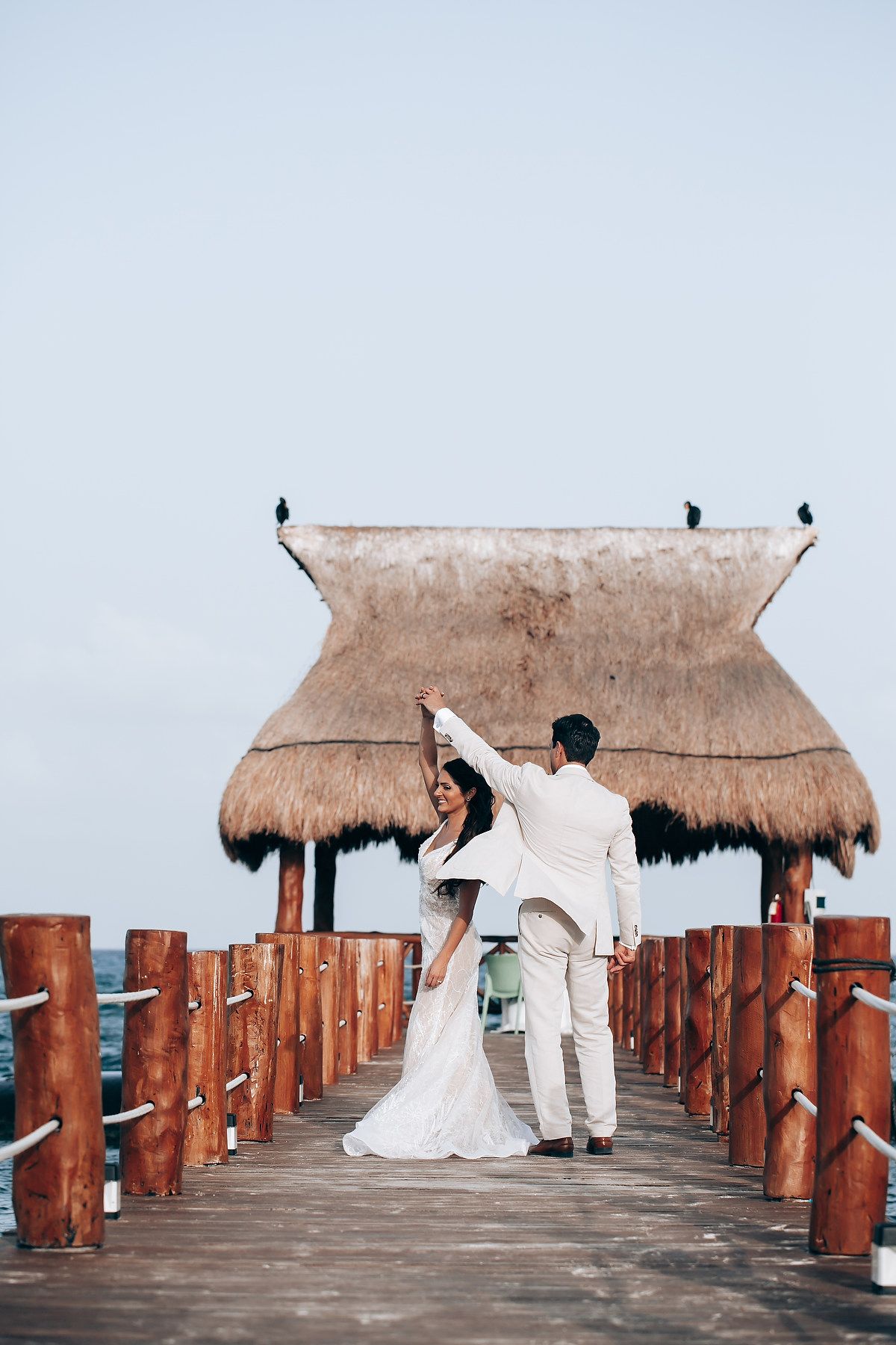 Newlyweds posing on a wooden boardwalk with thatched roof structures.