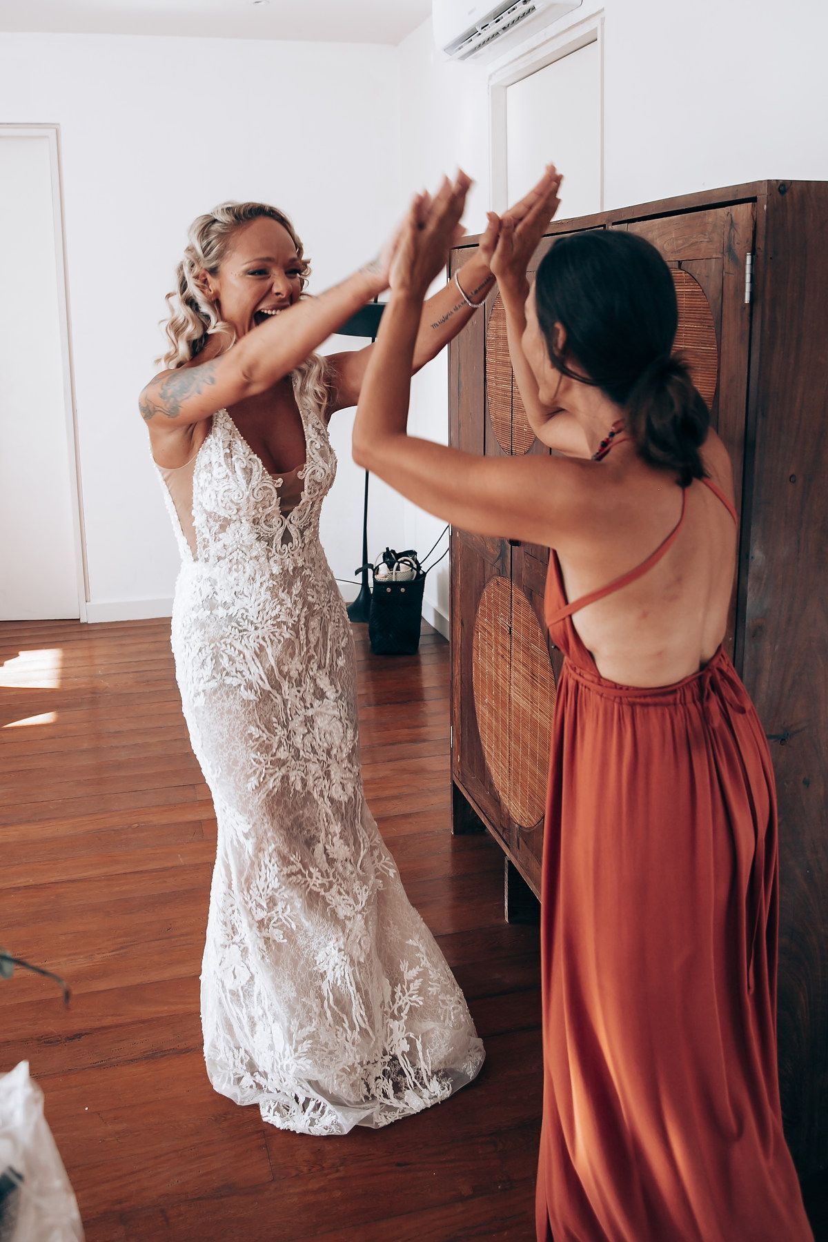 Two women dancing joyfully in a room with hardwood floors, one wearing a white lace dress and one in a rust-colored dress.