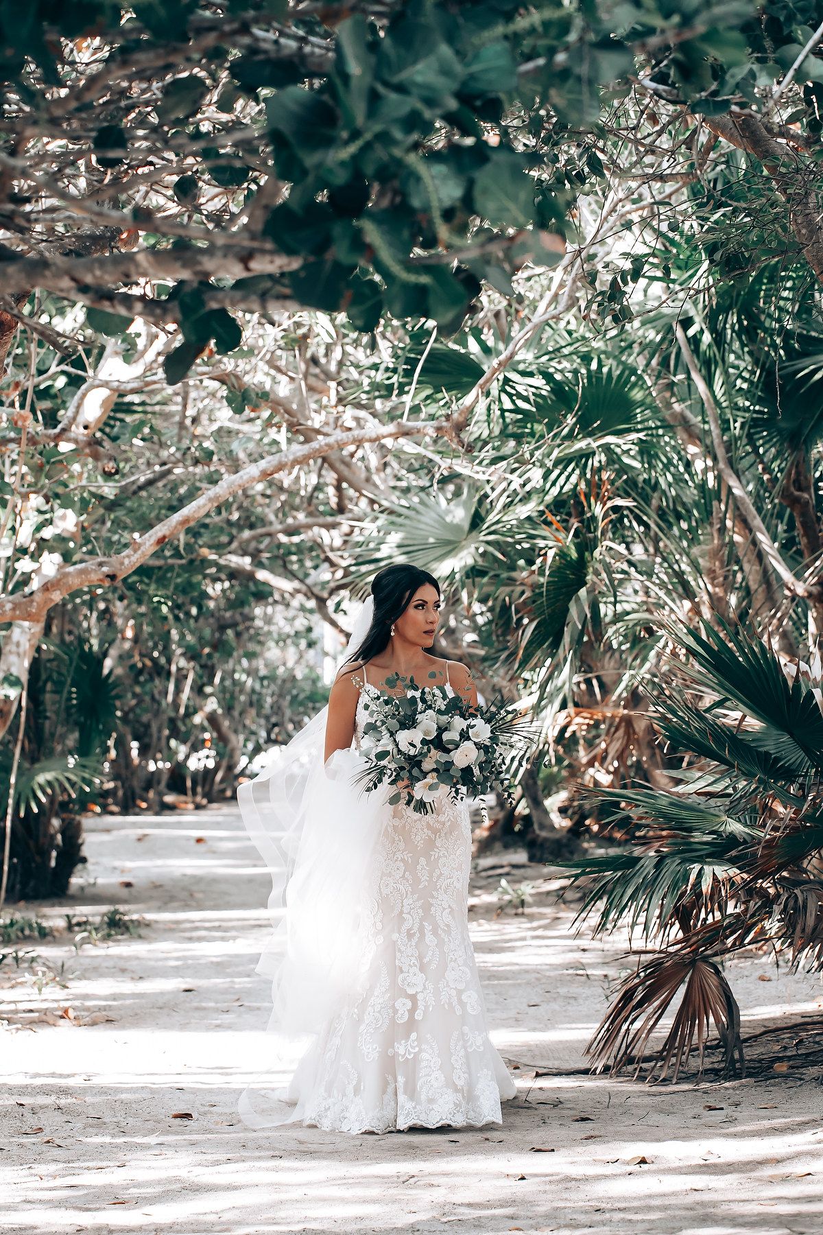 A white wedding dress flows elegantly against a tropical garden backdrop with palm trees and greenery.