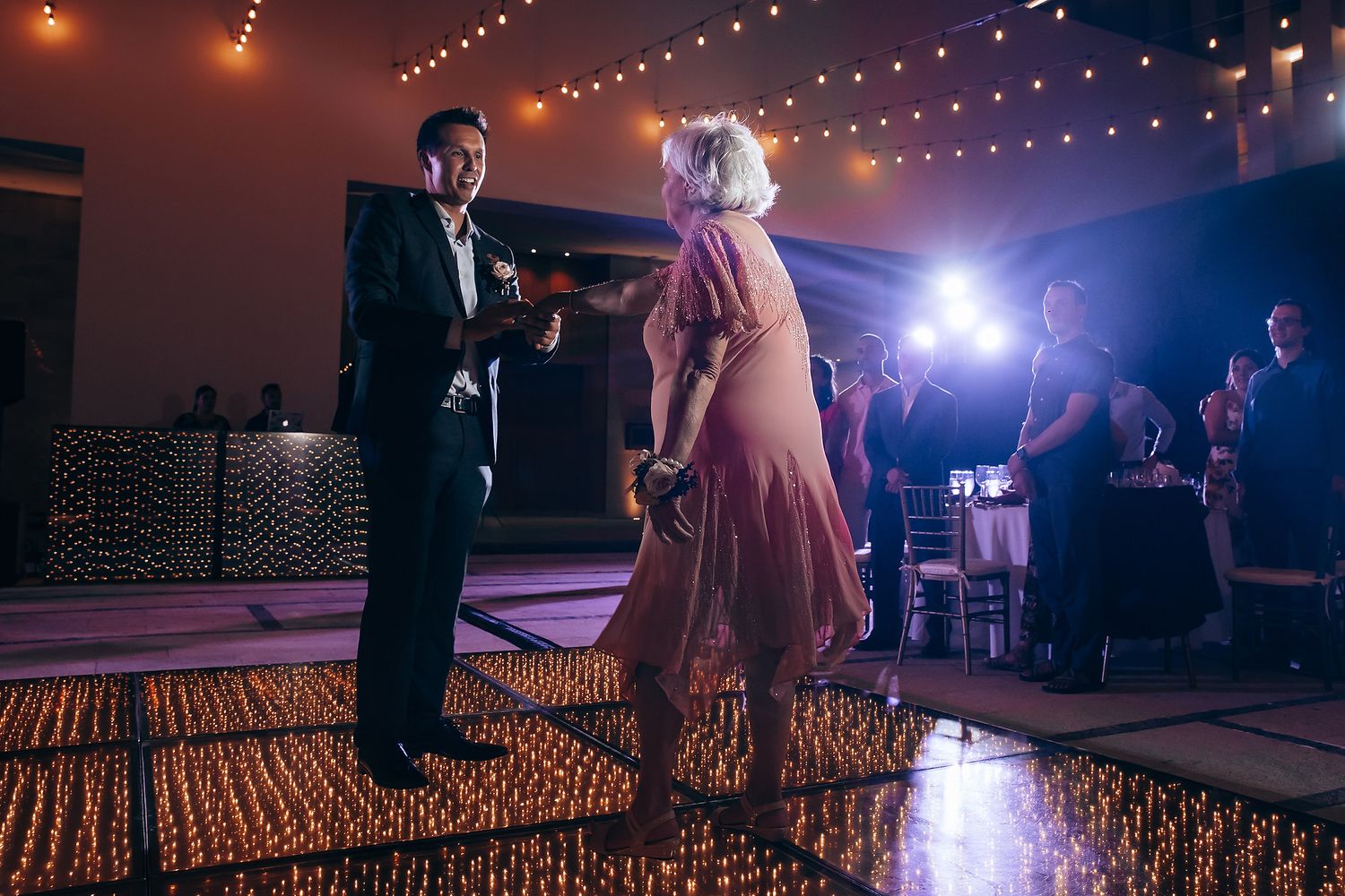 A couple dances together on a dimly lit dance floor with string lights overhead during an evening celebration.