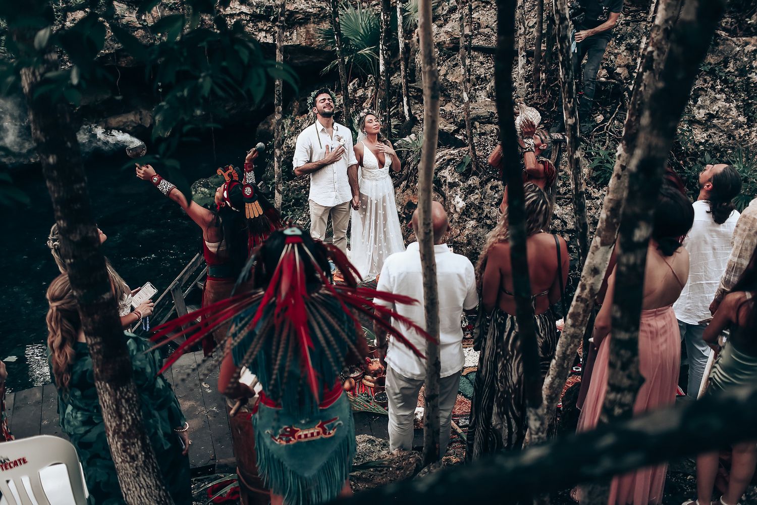 Guests in colorful attire celebrate at a wedding ceremony among trees and natural rock formations.