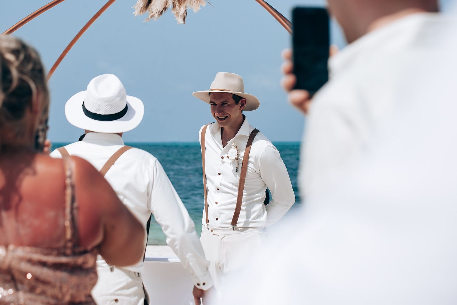 Elegant beach wedding celebration with attendees in white attire against turquoise ocean backdrop.