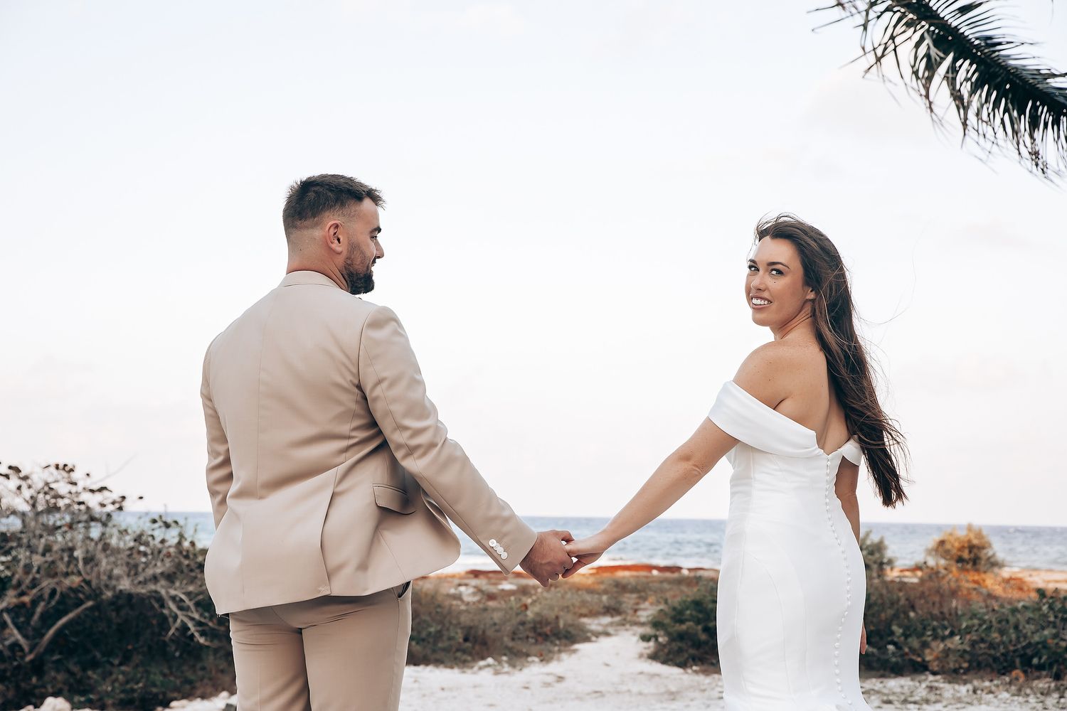 A couple in white and beige formal attire walking along a scenic coastal path during sunset.