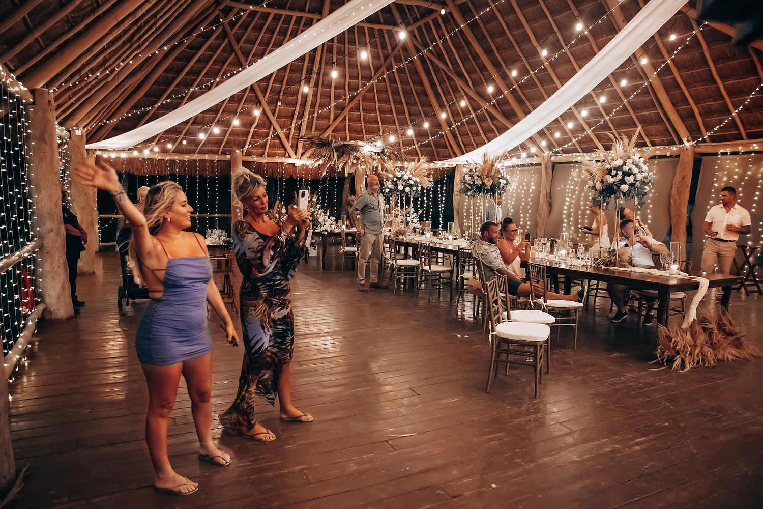 Wedding guests dancing together on an illuminated dance floor under twinkling string lights in a rustic barn venue.