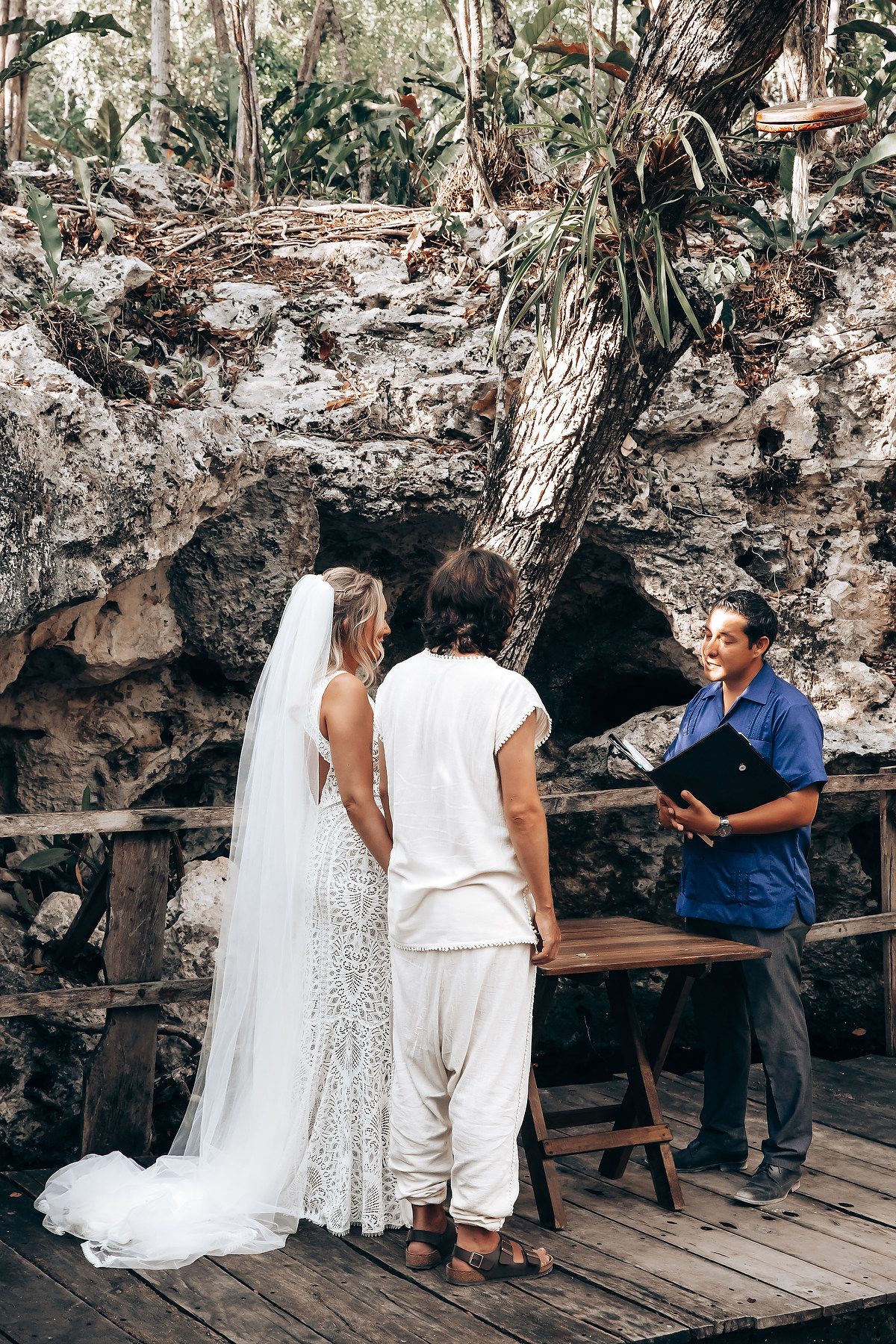 A couple in white outfits poses for photos on a wooden deck surrounded by rocky cliffs and tropical foliage.