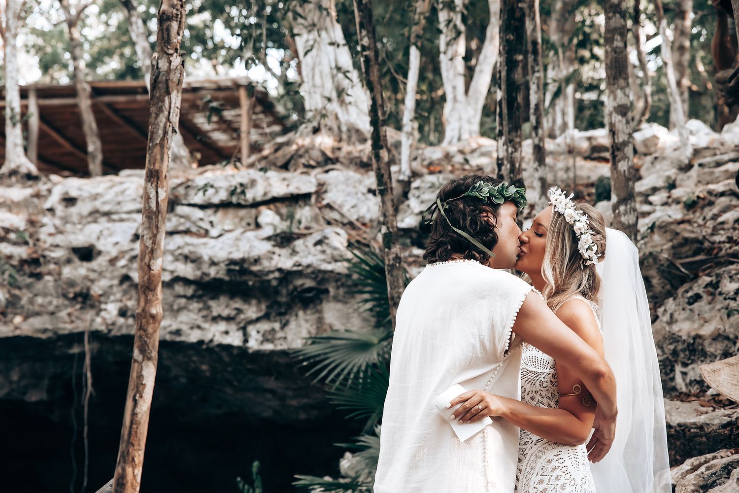 Couple in white attire poses intimately among tropical trees and foliage during outdoor photo session.