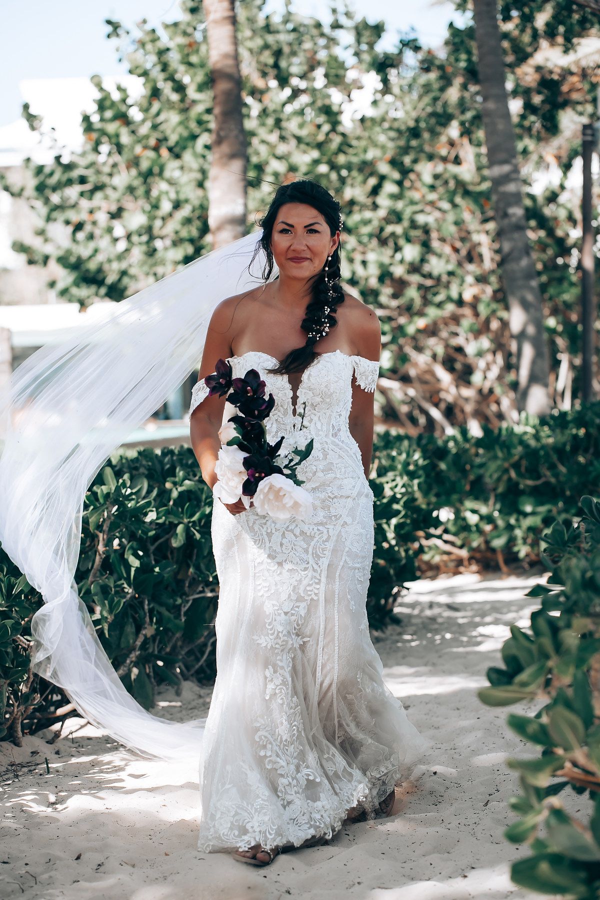 A flowing white wedding gown catches the breeze on a sandy tropical beach path.