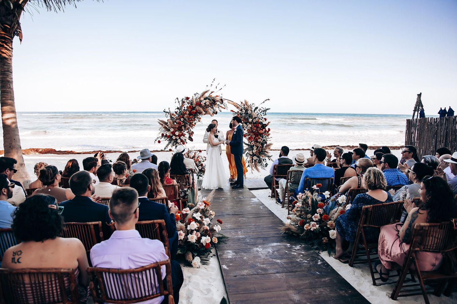 Beachfront wedding ceremony with guests seated under a floral arch overlooking the ocean at sunset.