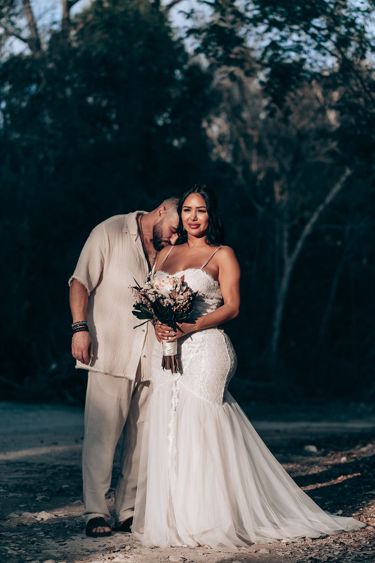 Wedding couple in white dress and beige suit embracing during an outdoor forest photoshoot.