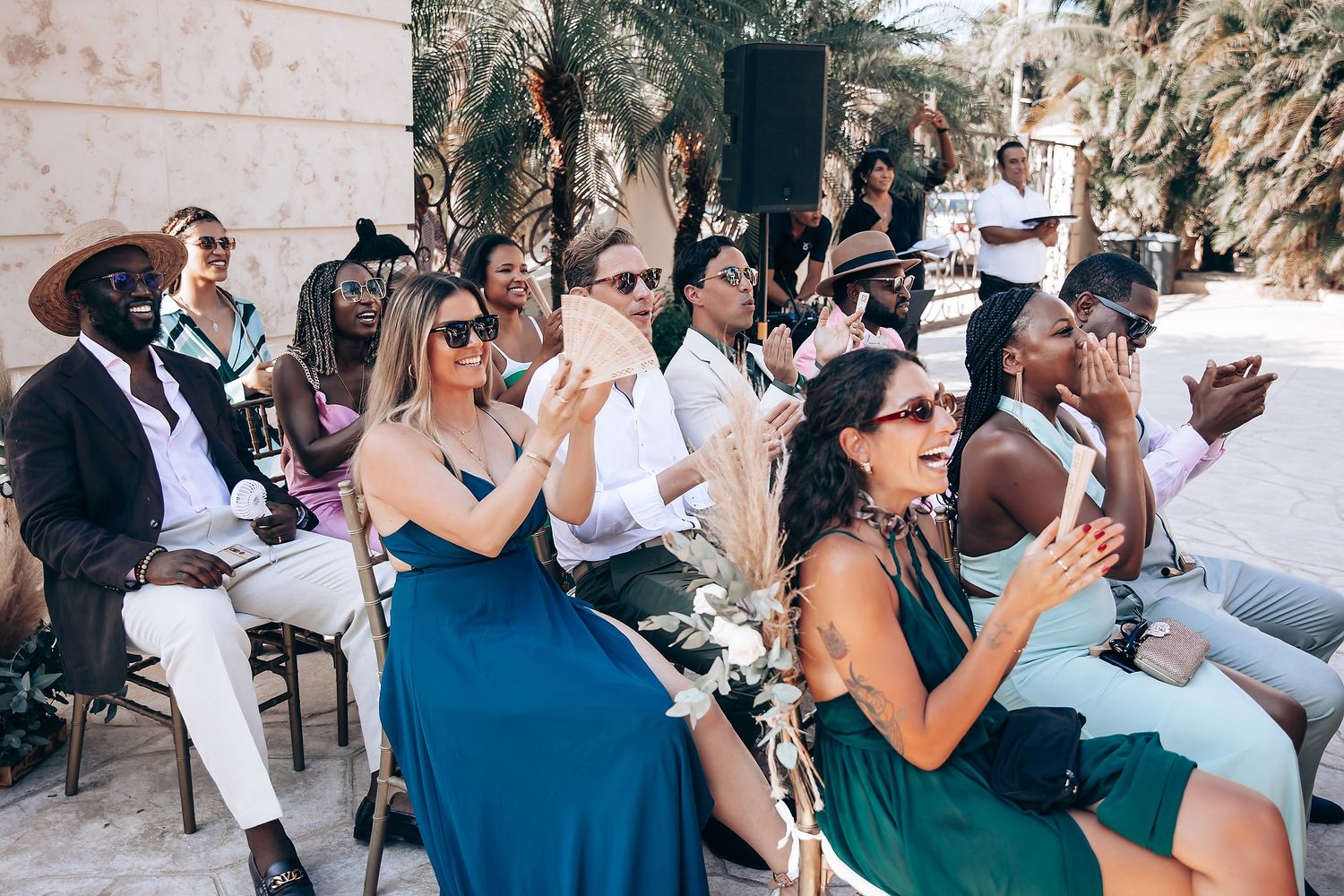 Wedding guests seated outdoors at a destination ceremony with tropical landscaping in the background.