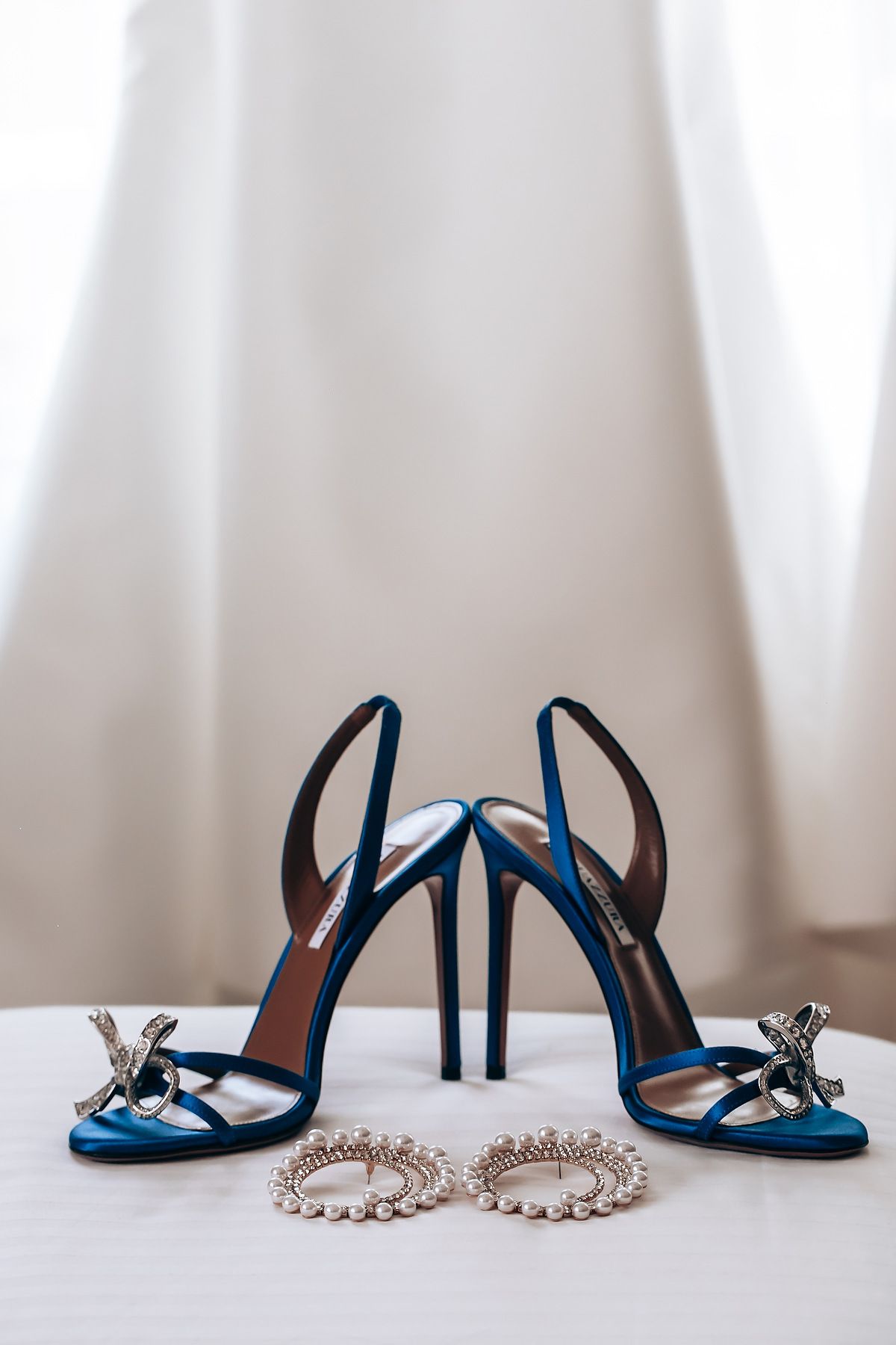 Elegant blue wedding heels with crystal embellishments and a pearl necklace displayed on white surface.