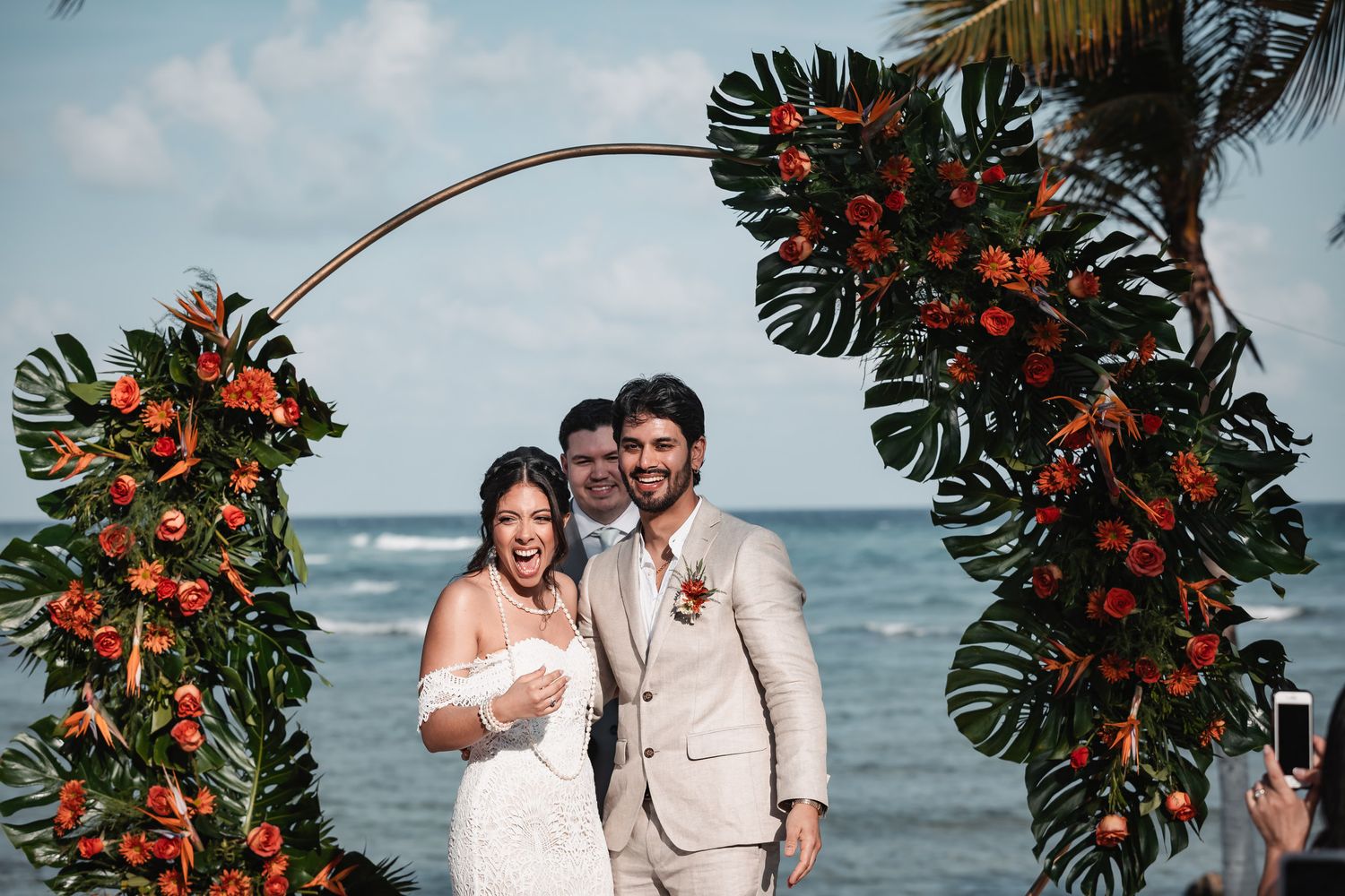 Newlyweds celebrate joyfully under floral arch at beachside wedding ceremony with ocean backdrop.