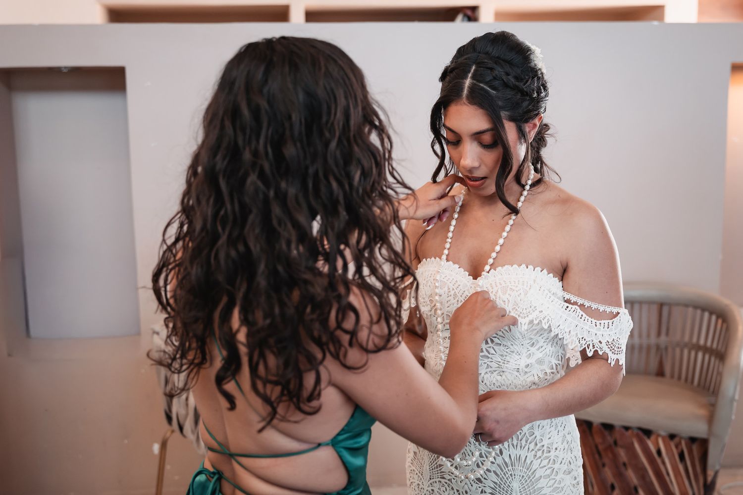 Bridesmaid in green dress helping bride in beaded off-shoulder wedding gown with final touch-ups before ceremony.