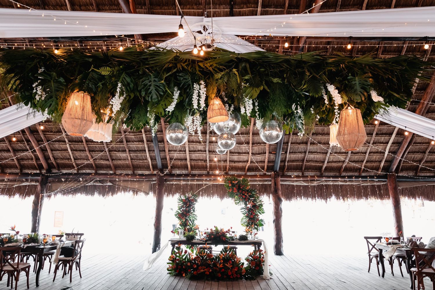 Tropical beach wedding reception venue with hanging greenery, globe lights, and floral centerpiece.