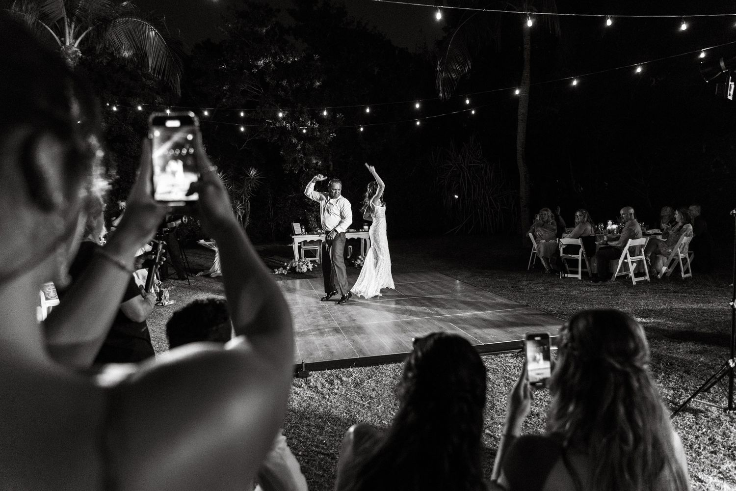 Black and white photo of wedding guests photographing couple's first dance on outdoor lit dance floor at night.