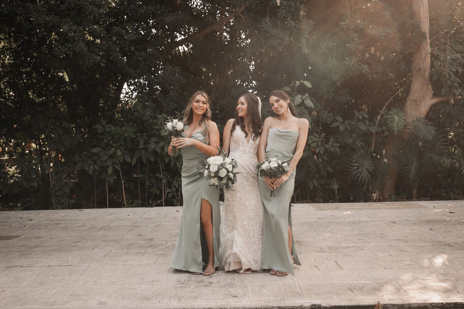 Bride in fitted lace gown poses with two bridesmaids in sage green dresses holding white bouquets outdoors.