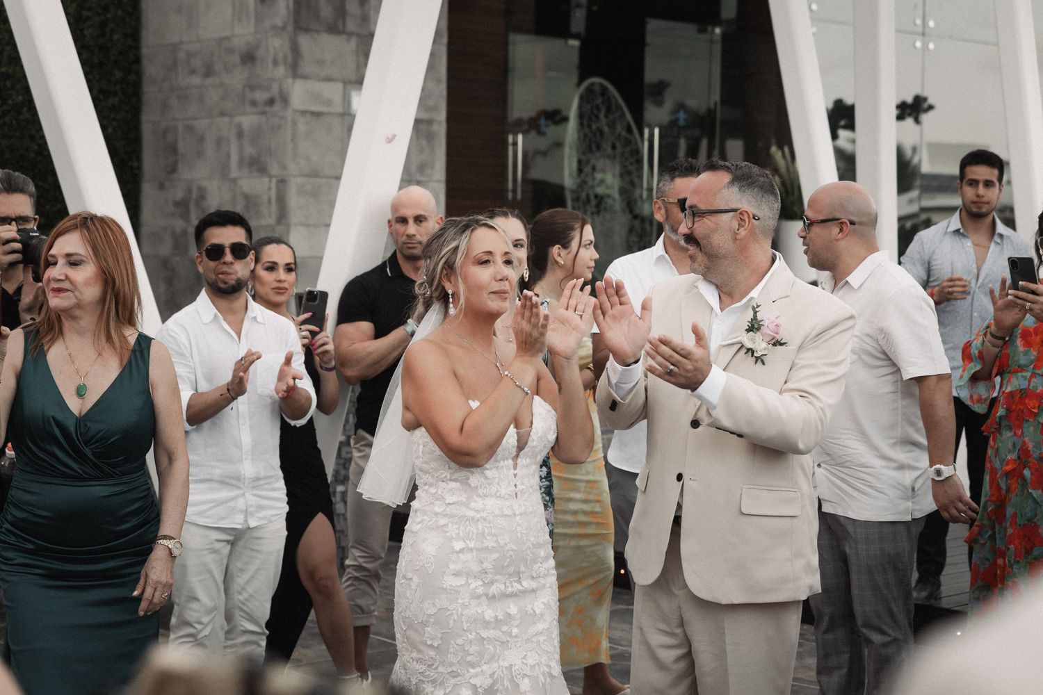 Bride in lace gown clapping and celebrating with wedding guests at outdoor reception venue.