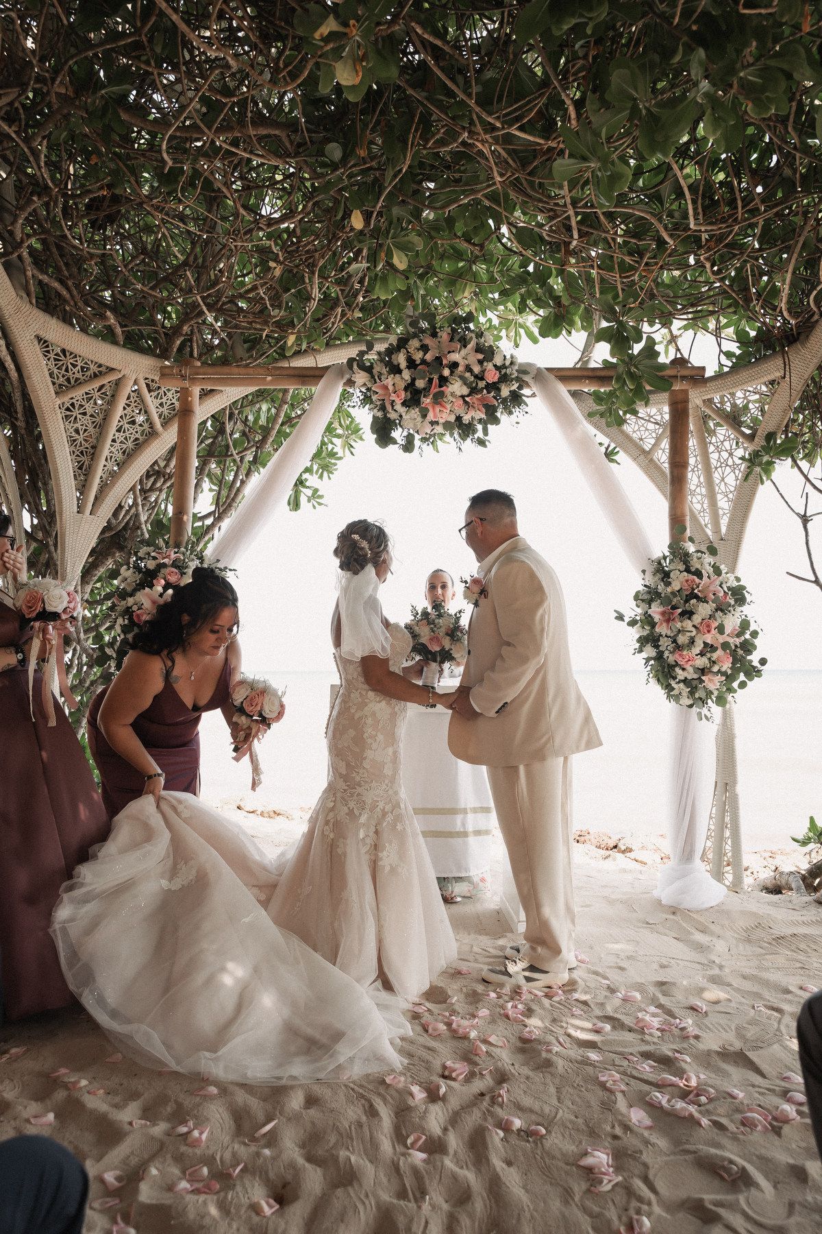 Bride and groom exchange vows under a floral arch on a sandy beach with a bridesmaid adjusting the wedding gown.