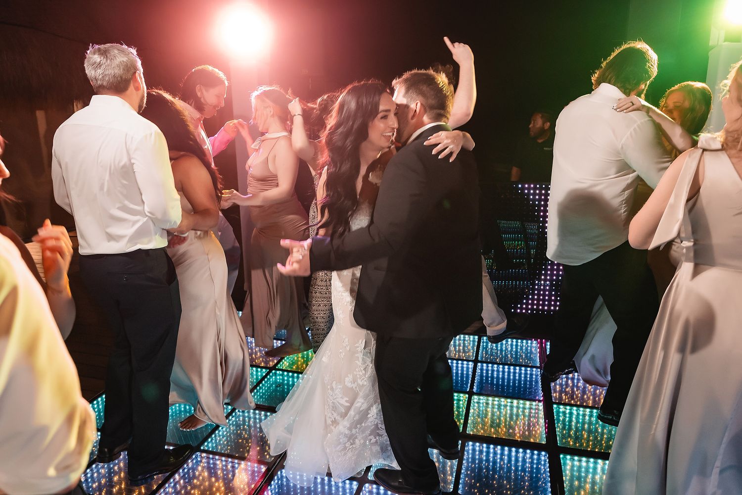 Bride and groom share a romantic dance on a colorful LED dance floor surrounded by wedding guests at a reception.