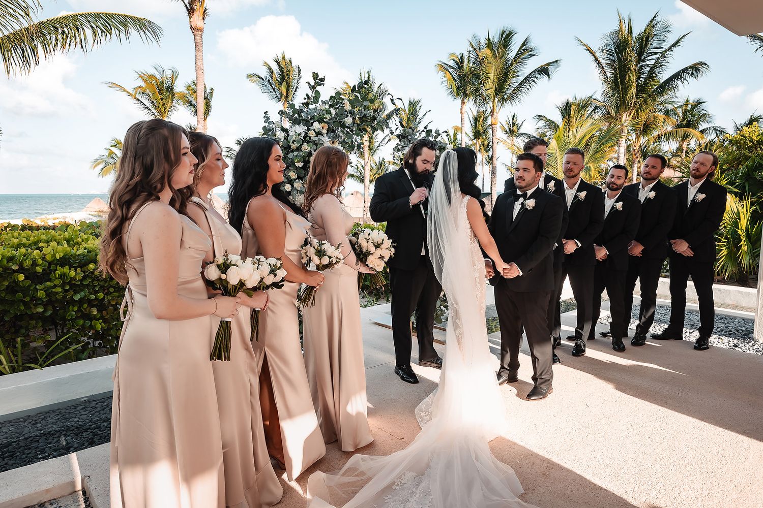 Bride and groom share a joyful moment with bridesmaids in champagne gowns and groomsmen at a tropical beachside wedding.