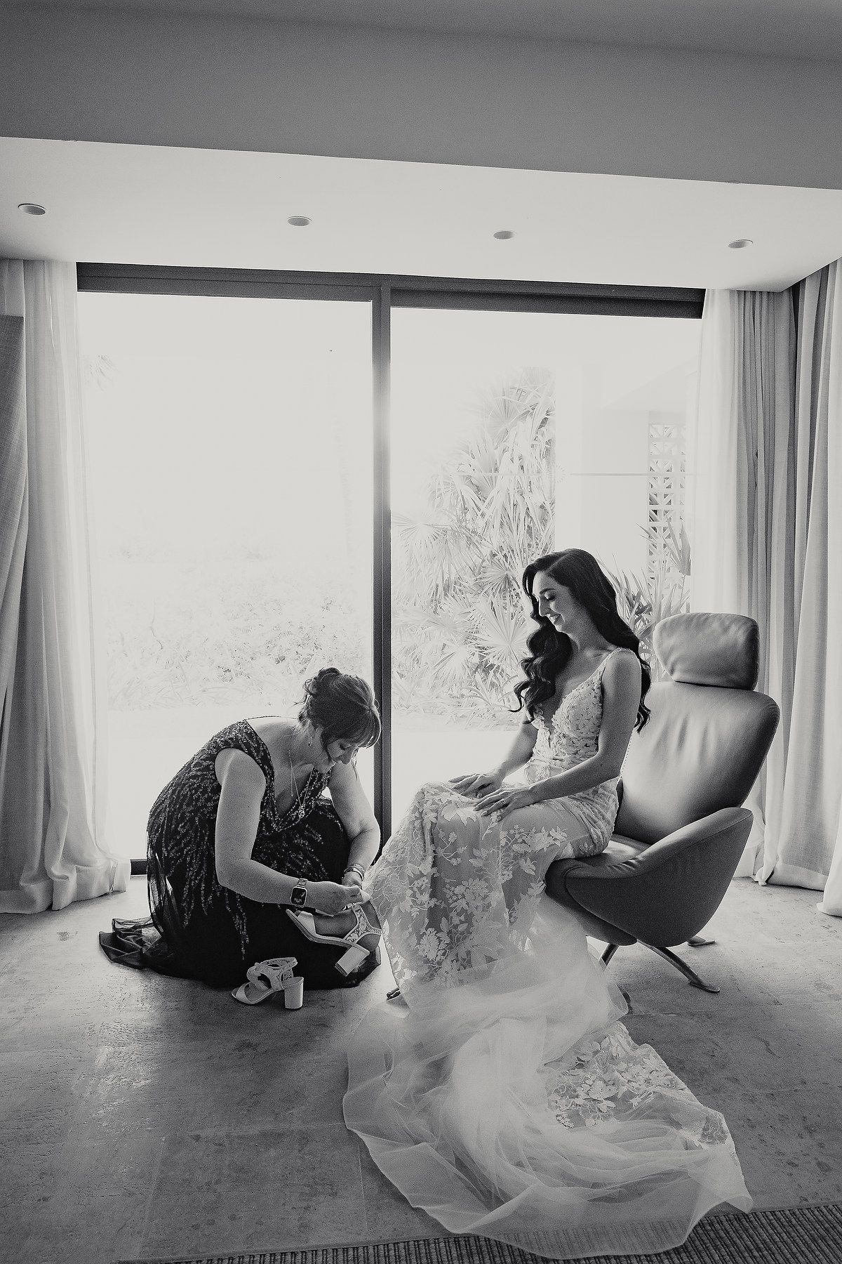 Black and white photo of a bride in a lace gown seated in a chair while an assistant helps with her shoes.