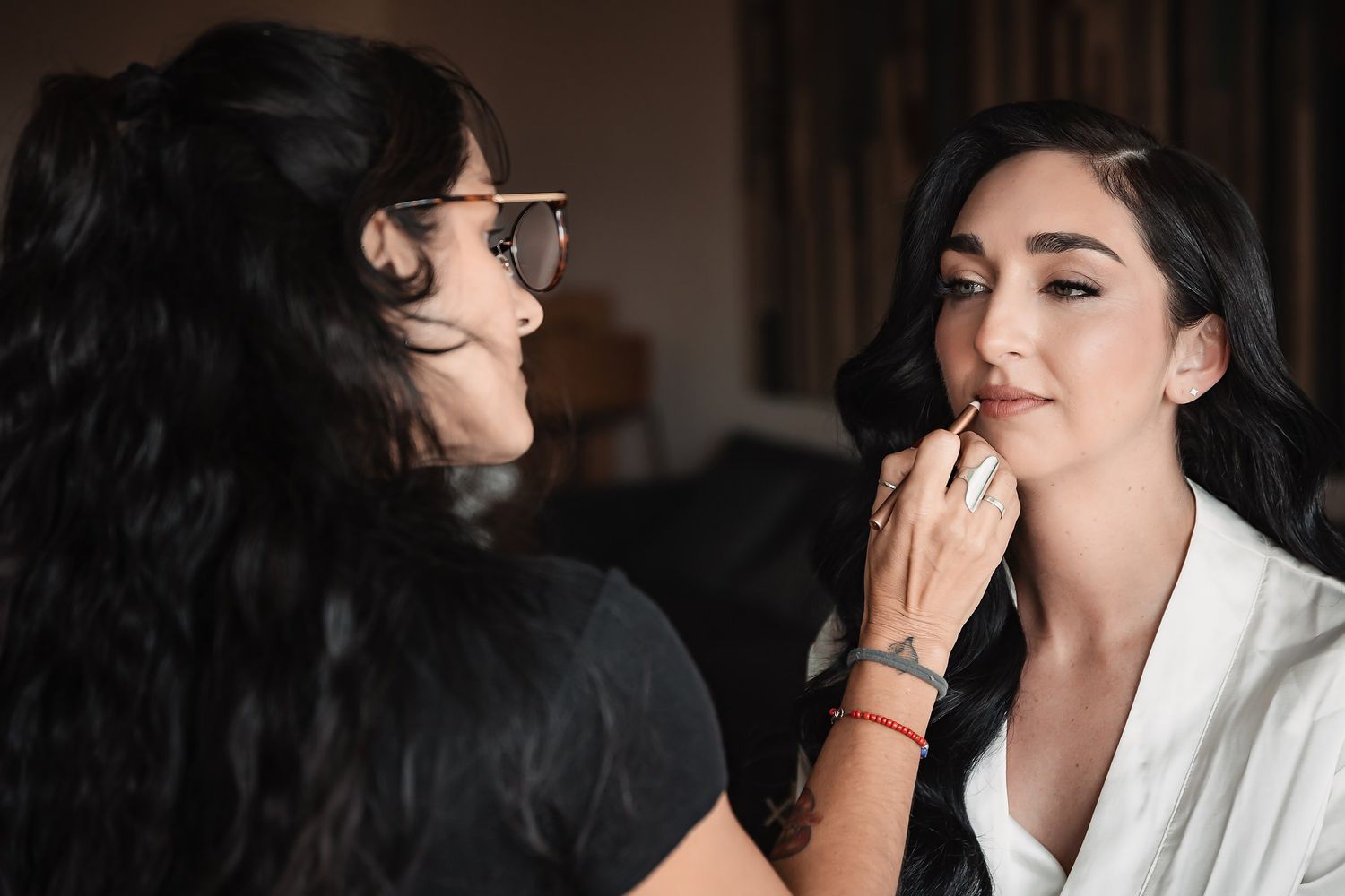 Makeup artist applying lipstick to brunette bride during wedding day preparations indoors.