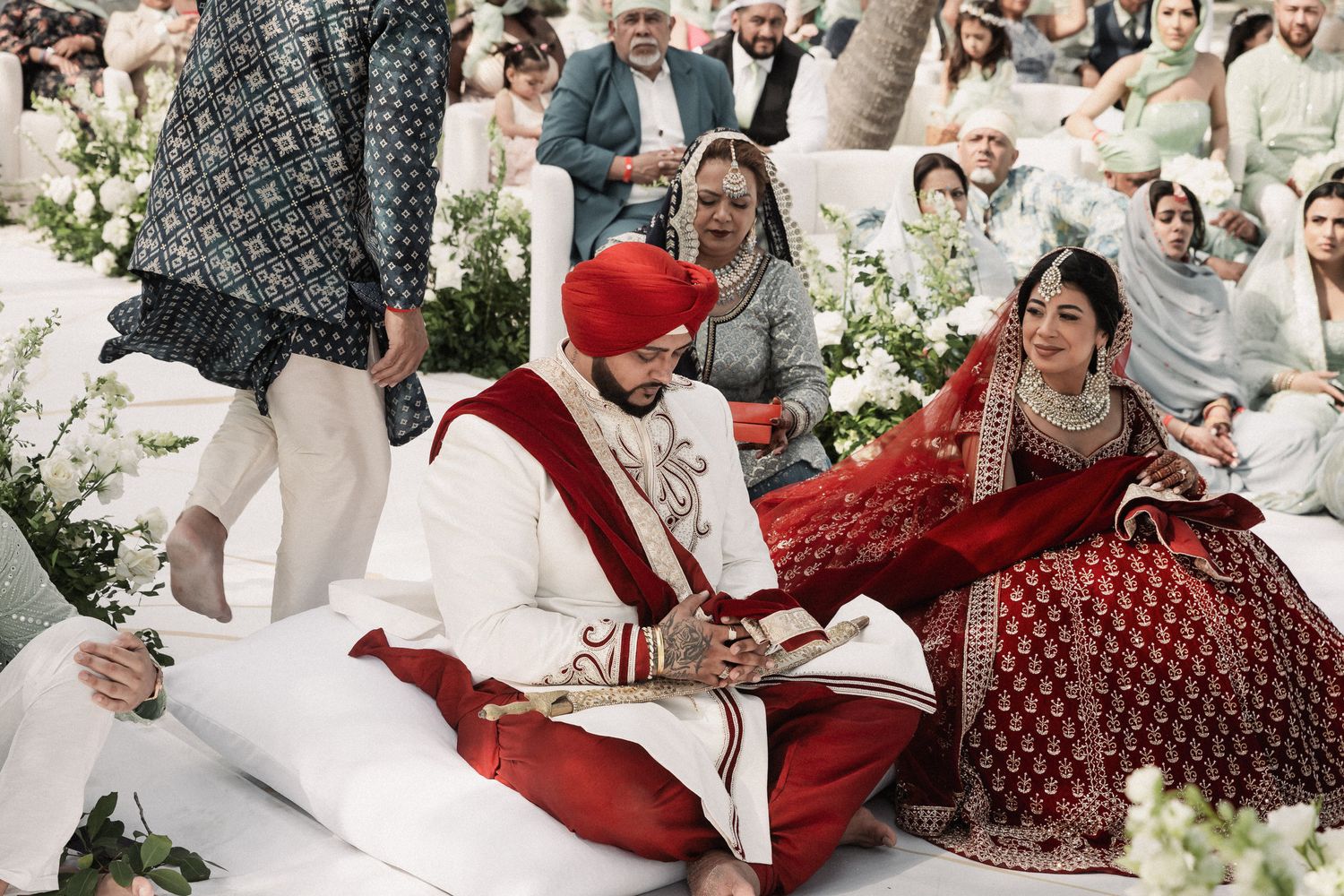 Indian bride in red lehenga and groom in white sherwani with red turban seated during traditional Sikh wedding ceremony outdoors.