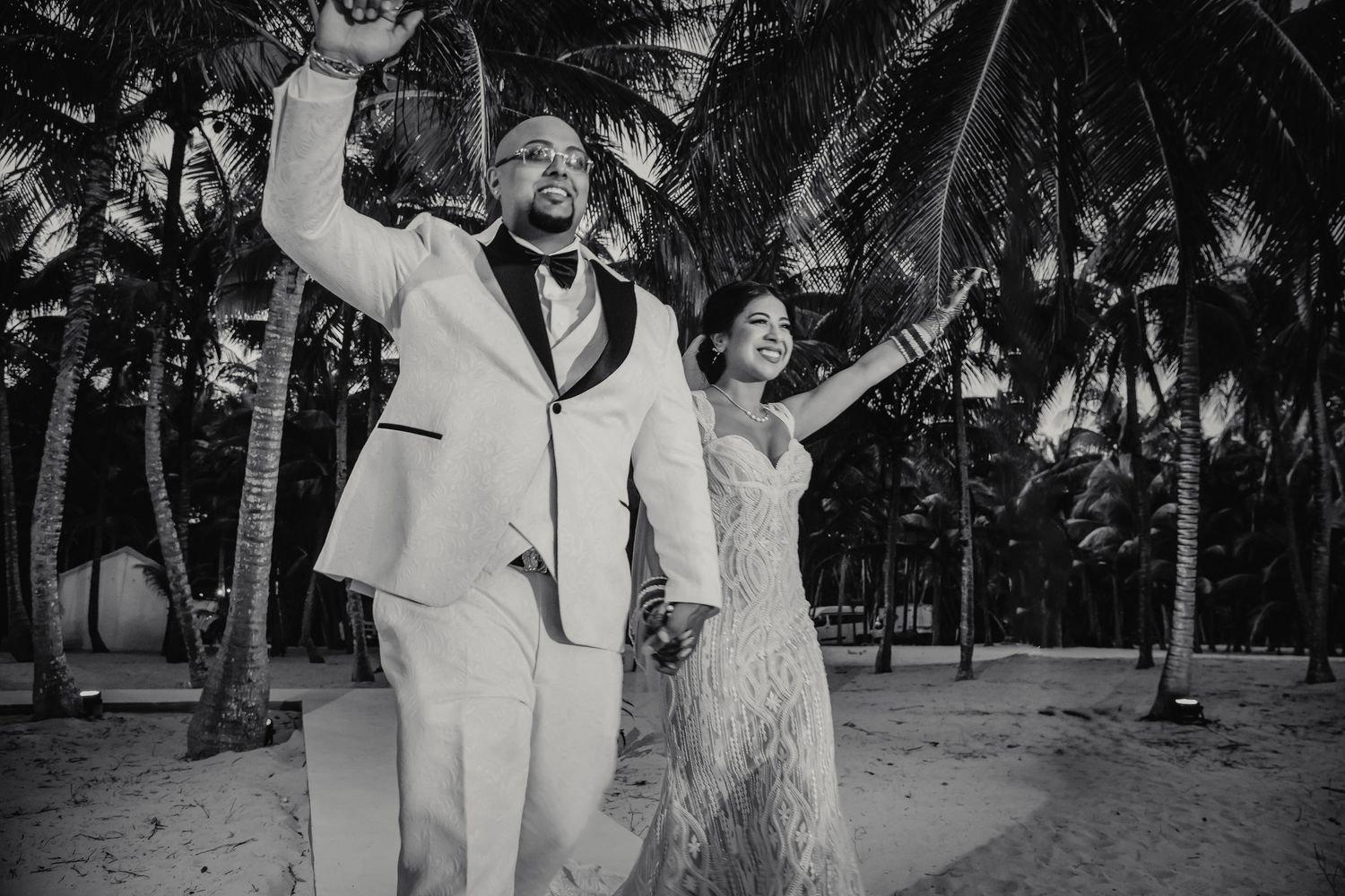 Joyful newlyweds celebrate on a tropical beach surrounded by palm trees in a stunning black and white wedding photo.