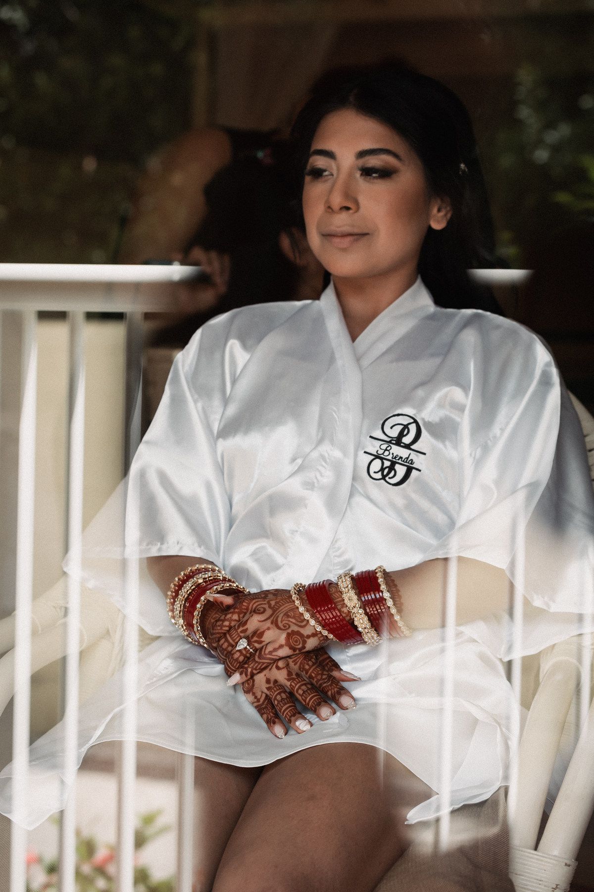 Bride in white satin robe with mehndi henna hands and red bangles sitting before Indian wedding ceremony.