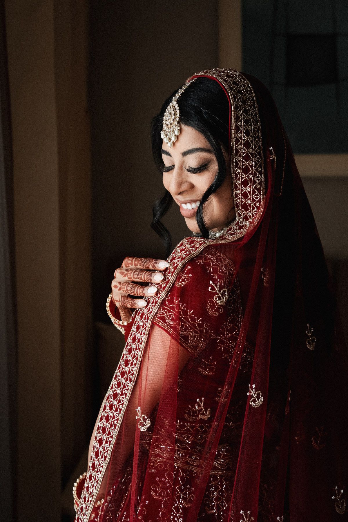 Beautiful Indian bride in red lehenga with gold embroidery and dupatta smiling softly indoors.
