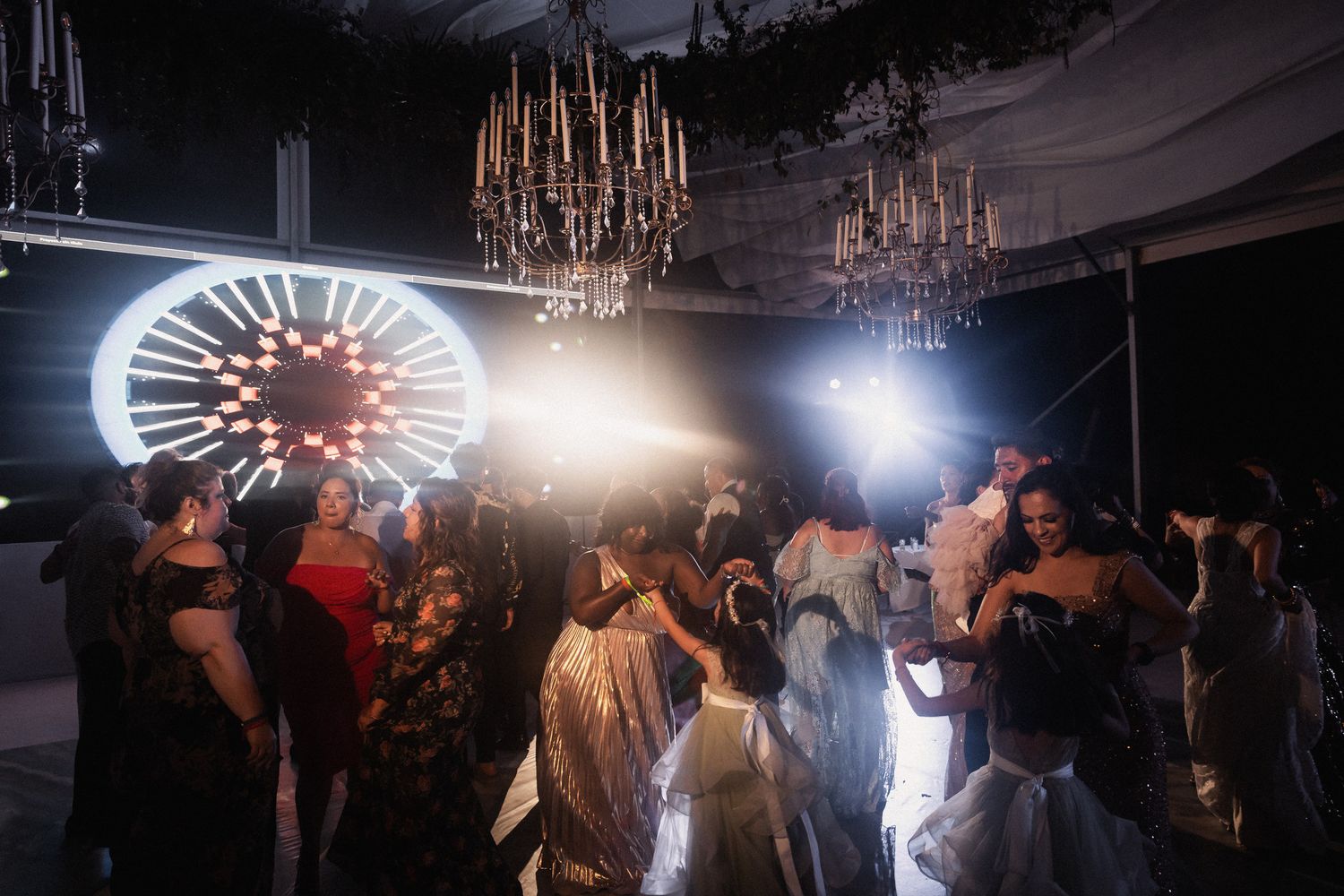 Guests dancing under crystal chandeliers at an elegant evening wedding reception with dramatic lighting.