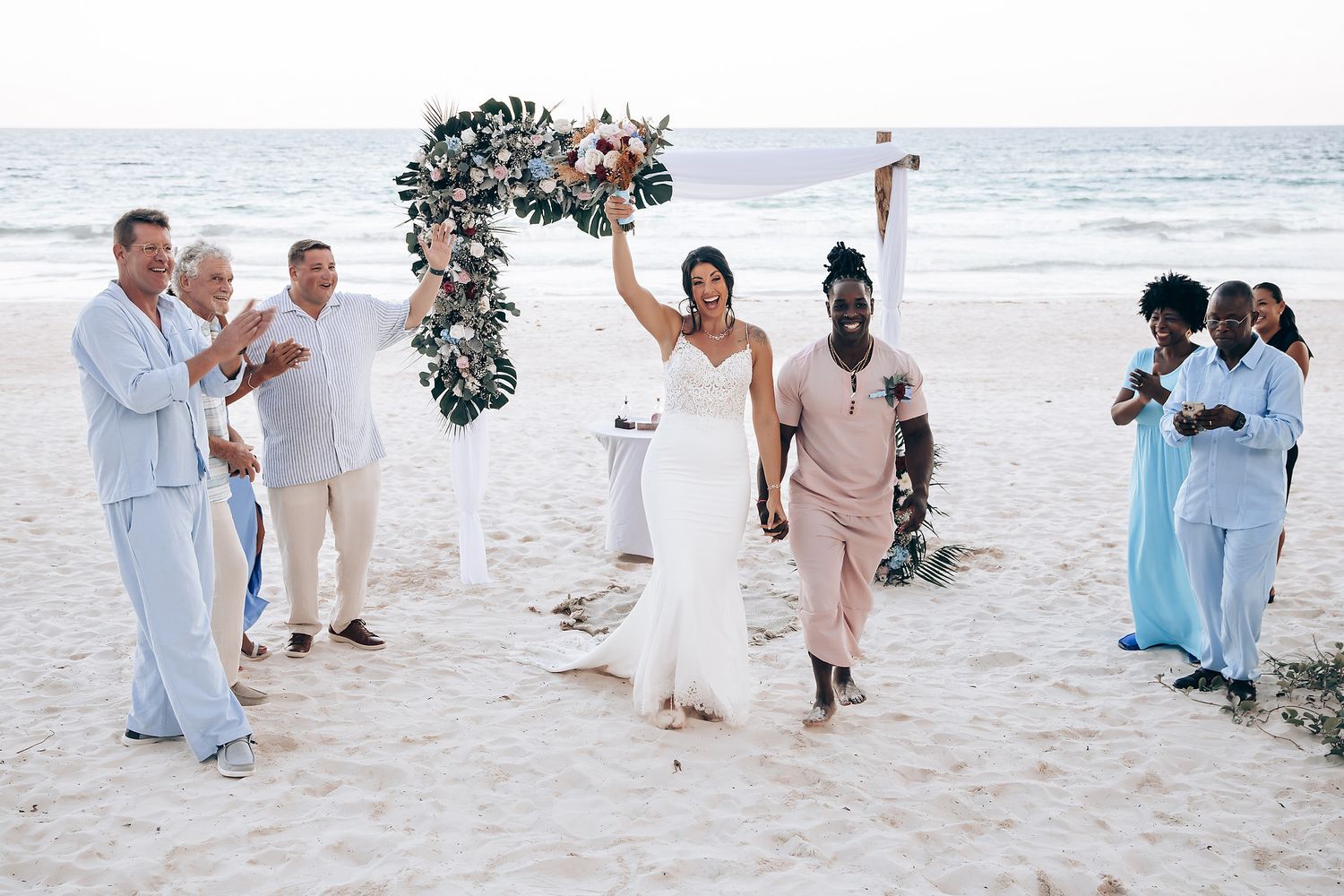 A wedding ceremony on a beach, with the couple and their wedding party standing in front of a floral arch.