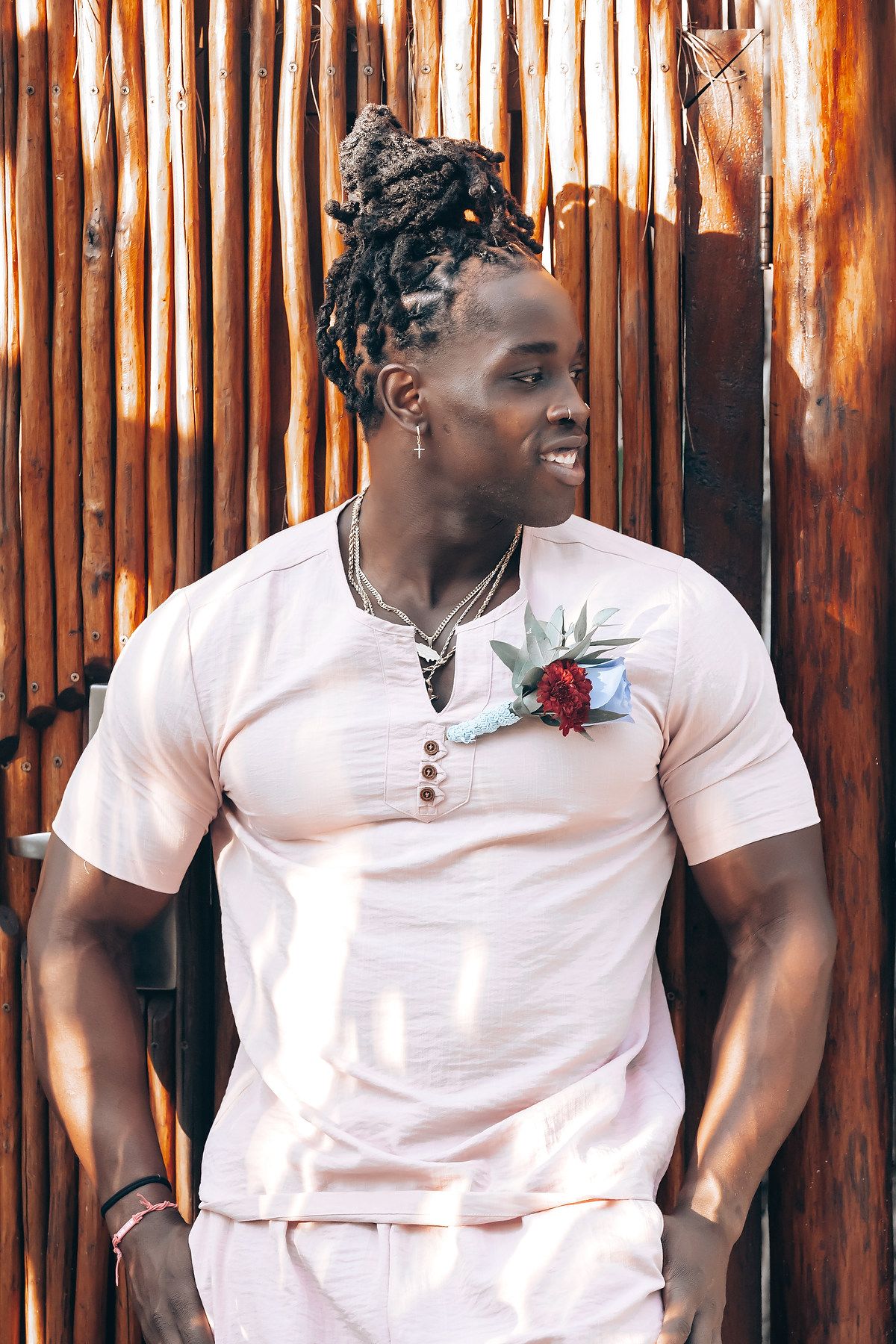 A man with dreadlocks wearing a white shirt stands in front of a wooden fence.