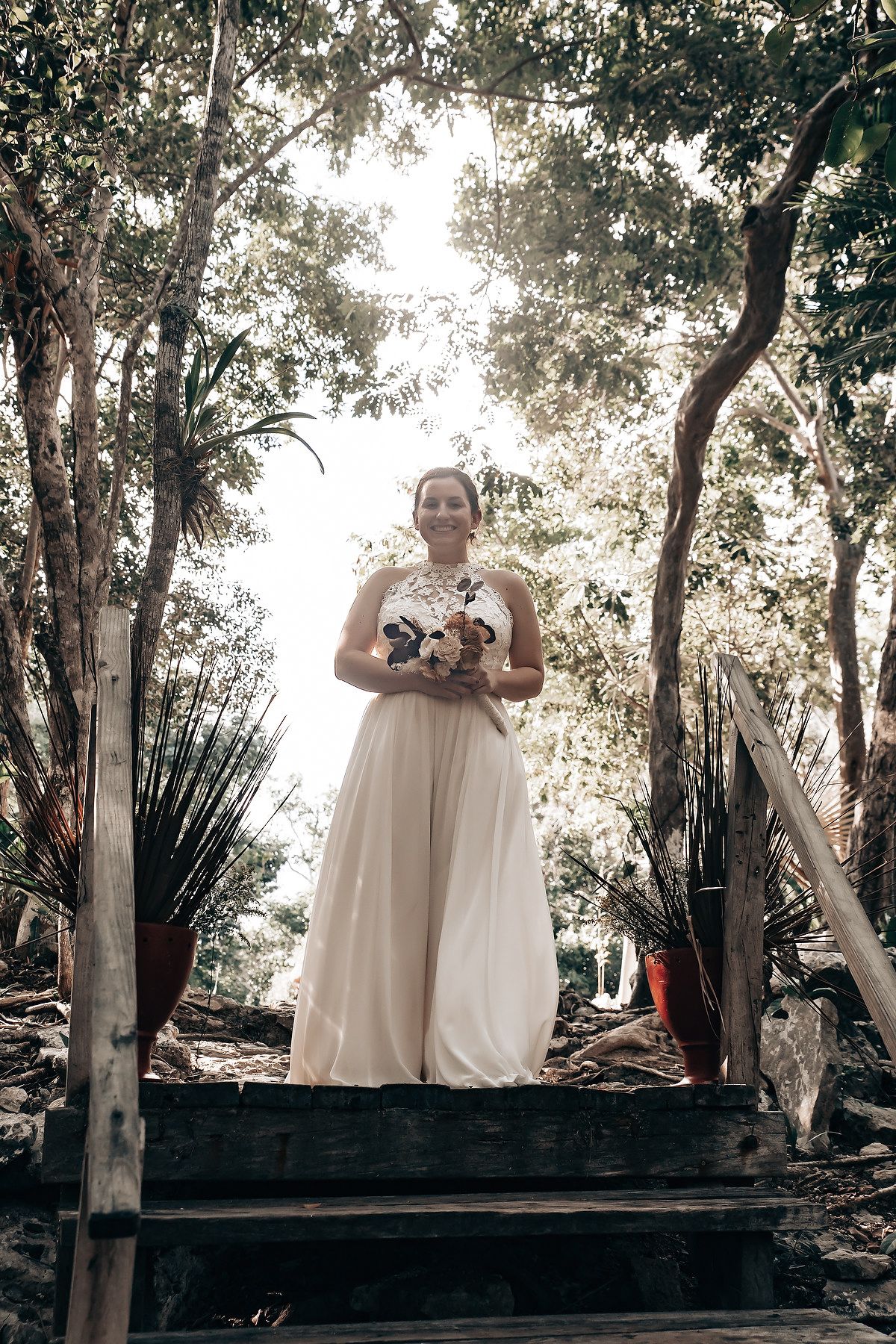 A woman in a white wedding dress standing on a wooden platform in a forest with trees and foliage in the background.