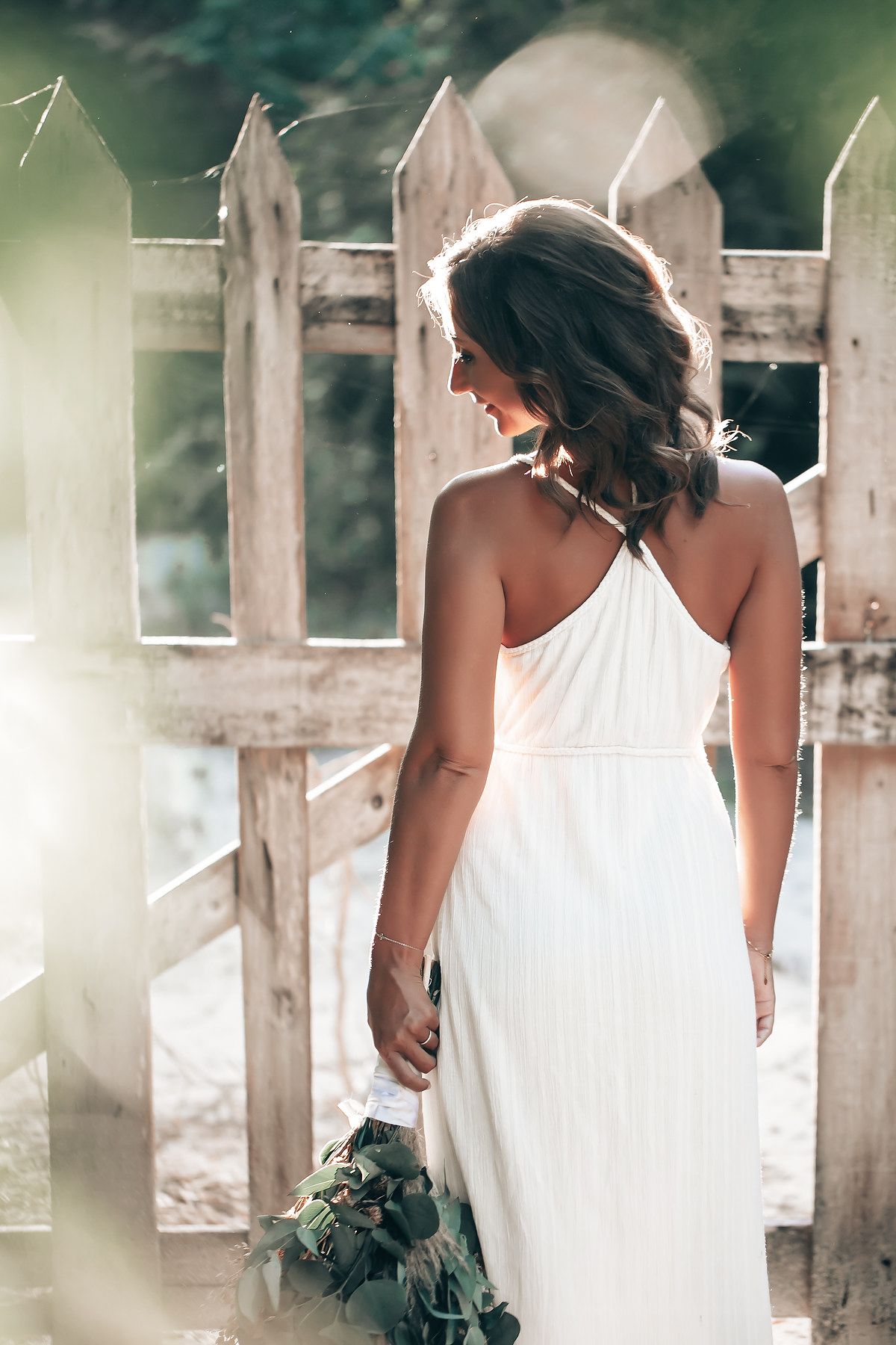 A woman in a white dress stands facing away from the camera in a forested area.