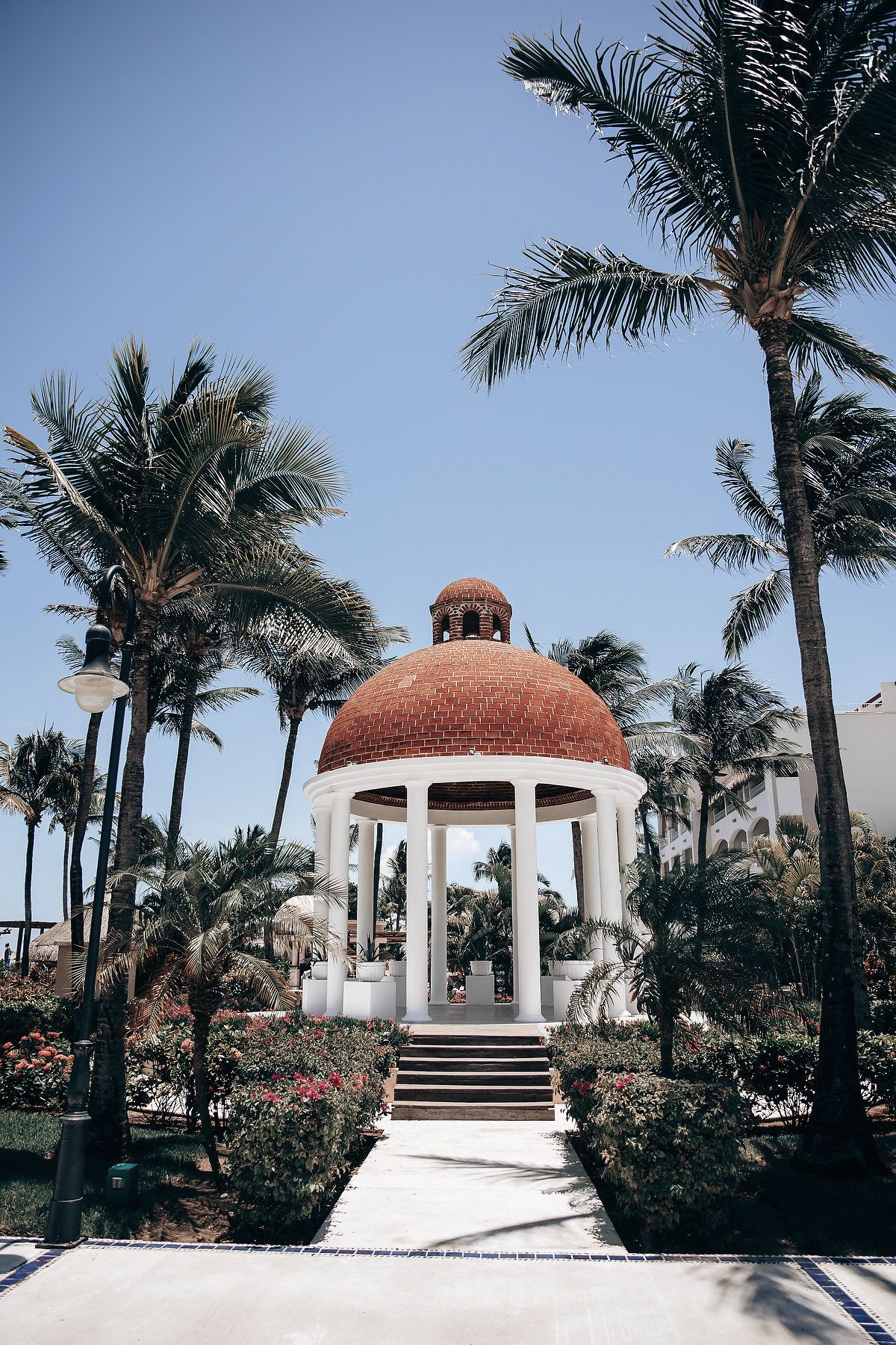 A white gazebo with a domed roof surrounded by palm trees against a clear blue sky.